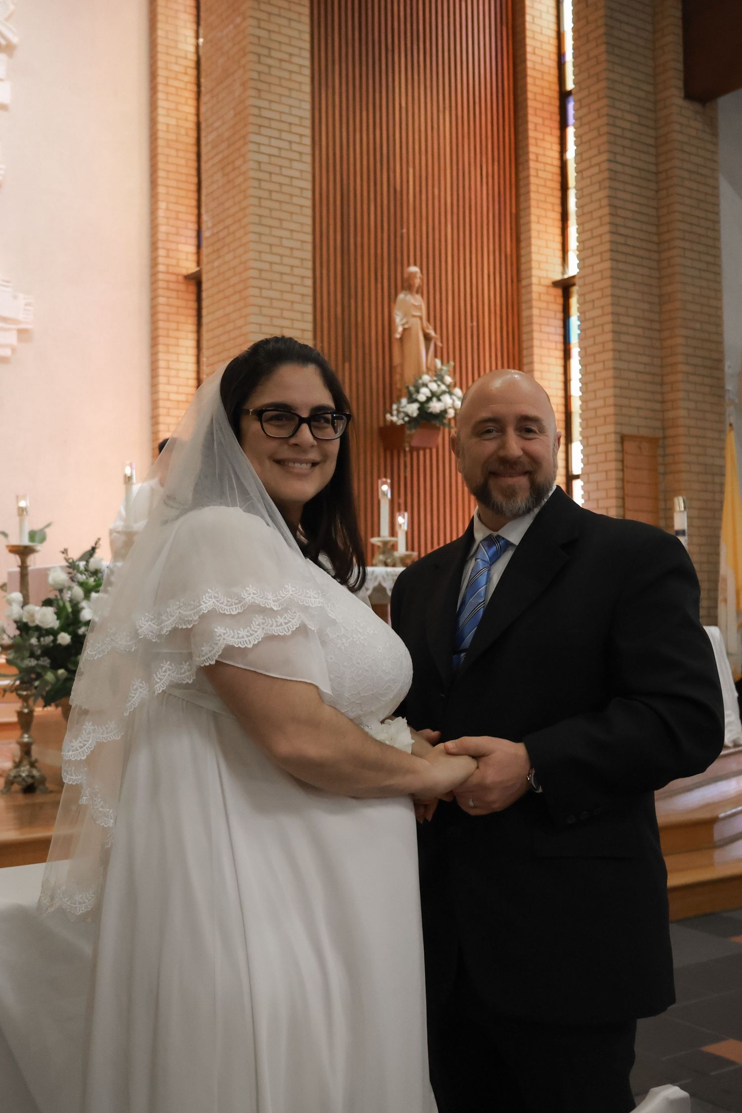 Smiling bride in white gown and veil with groom in black suit holding hands at church altar ceremony.