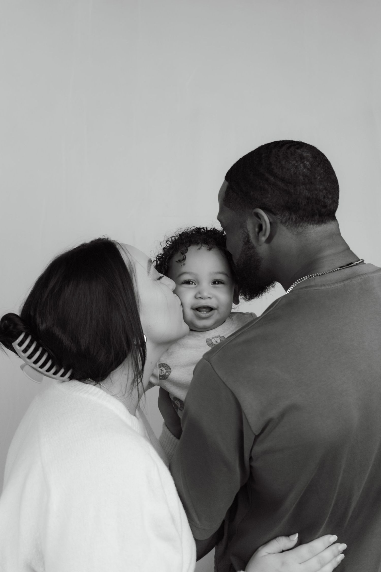 Black and white photo of interracial couple kissing their smiling baby together against a light background.
