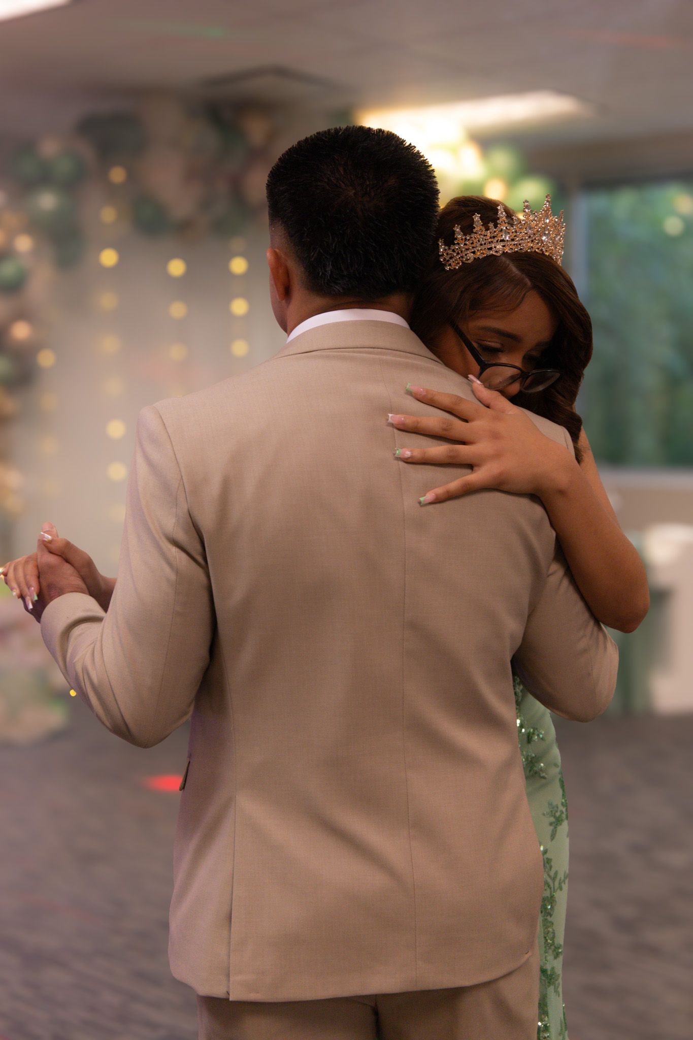 Father and daughter share an intimate dance the woman wearing a gold crown and green dress, embraced by a man in a beige suit.
