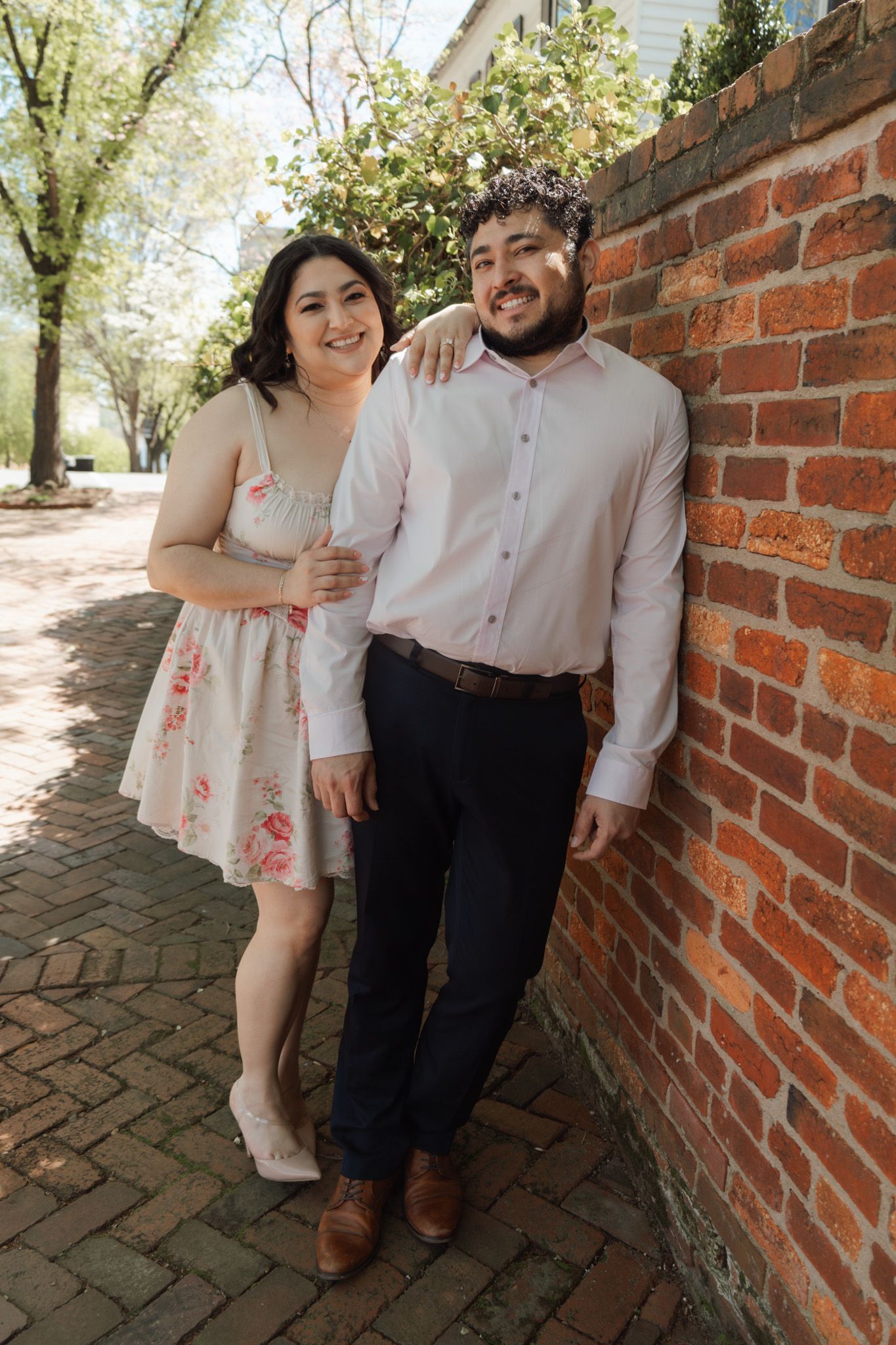 Smiling couple posing against a red brick wall outdoors, woman in floral dress and man in pink shirt, spring trees in background.