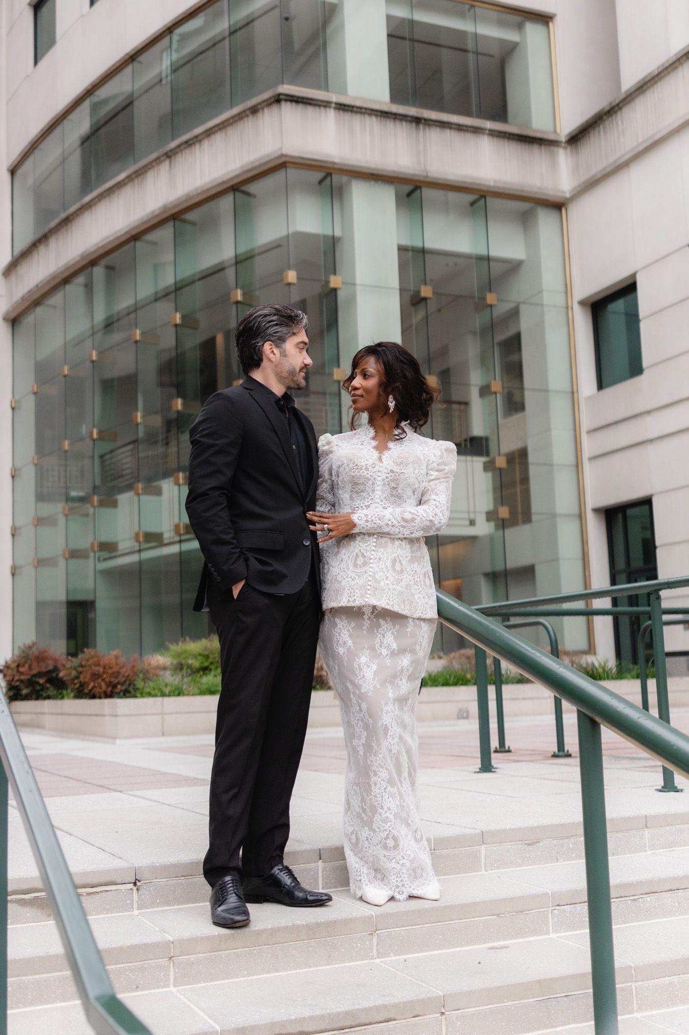 Elegant interracial couple in formal attire smiling on courthouse steps, man in black suit, woman in white lace gown elopement.