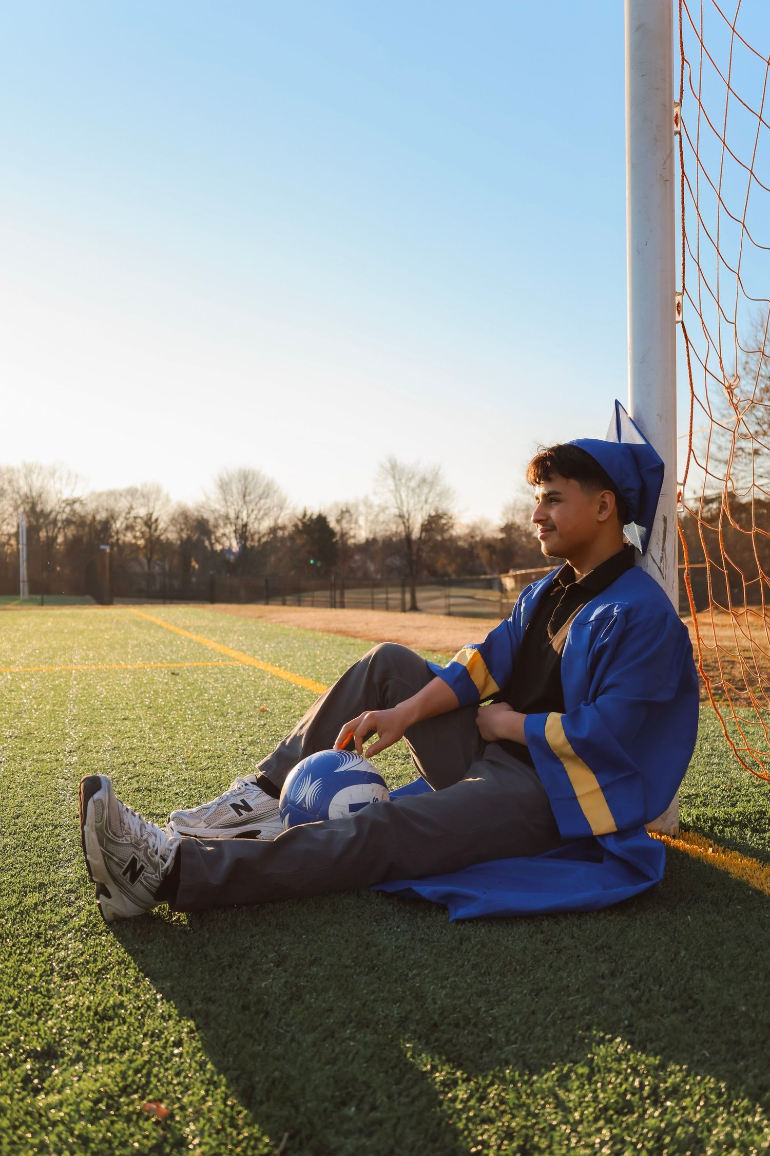 Young man in blue graduation cap and gown sitting on soccer field grass, leaning against goal post on sunny day.