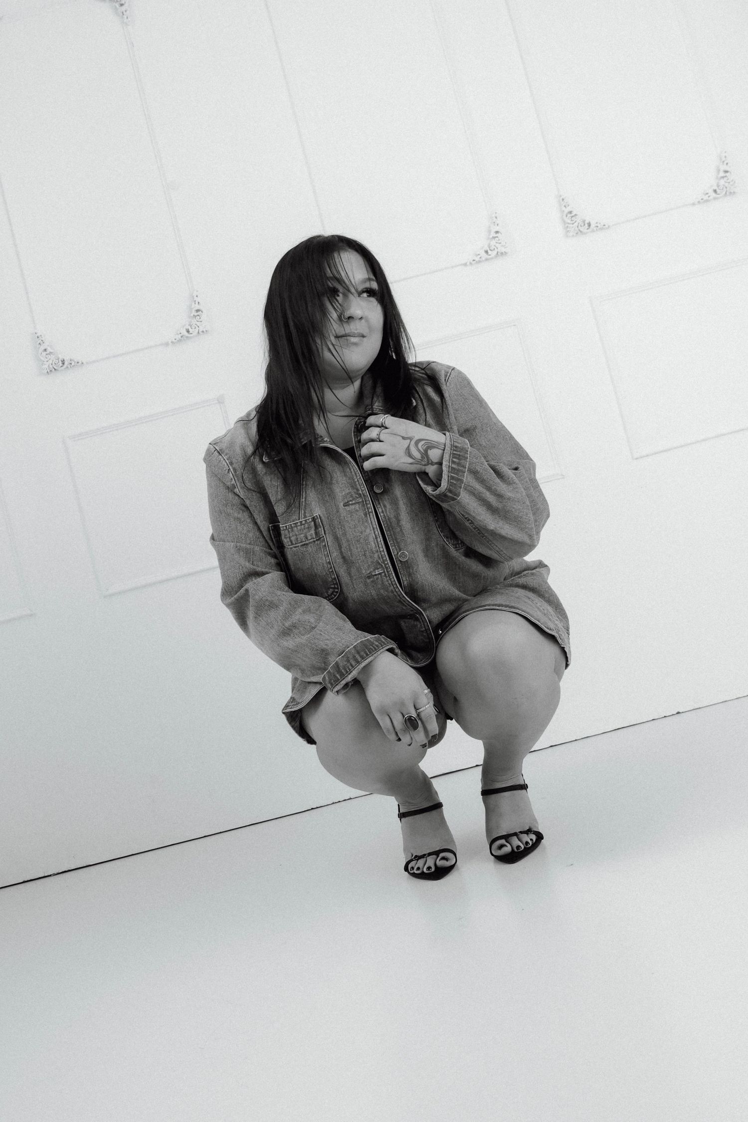 Black and white photo of a woman crouching in a denim jacket and heels against a white paneled wall in a studio.