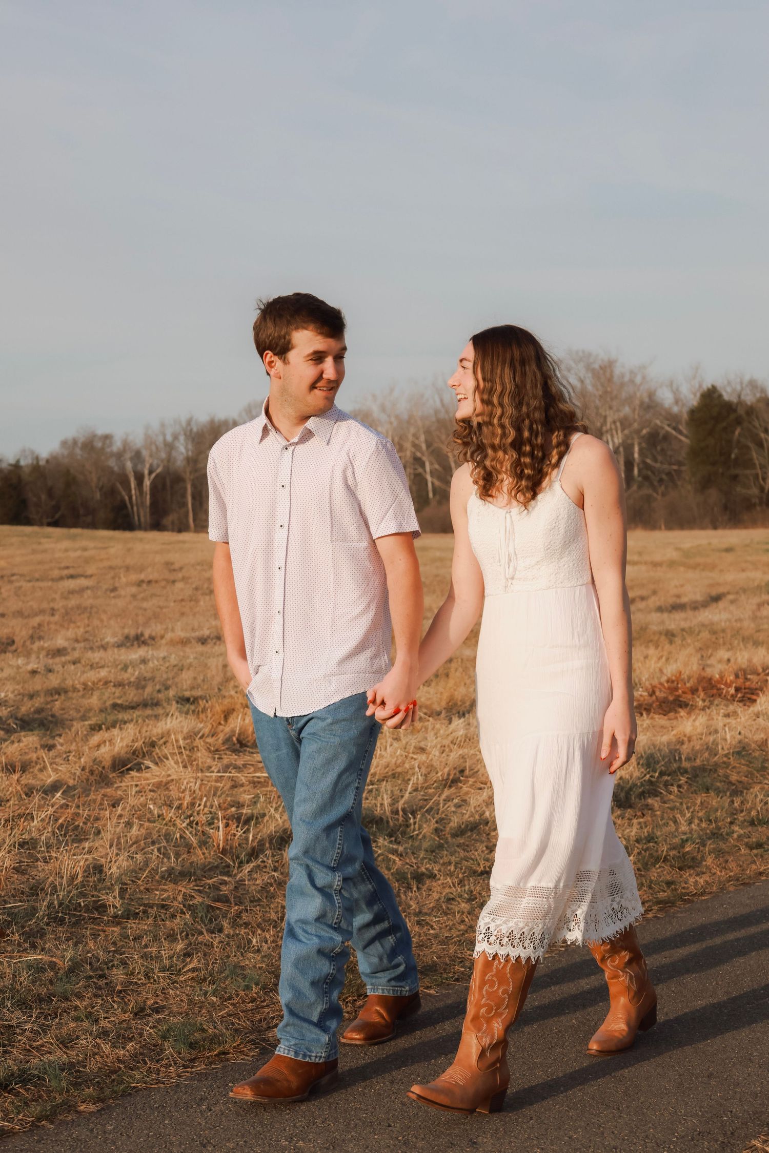 Young couple holding hands walking on a rural road, woman in white dress and cowboy boots, man in jeans and white shirt.