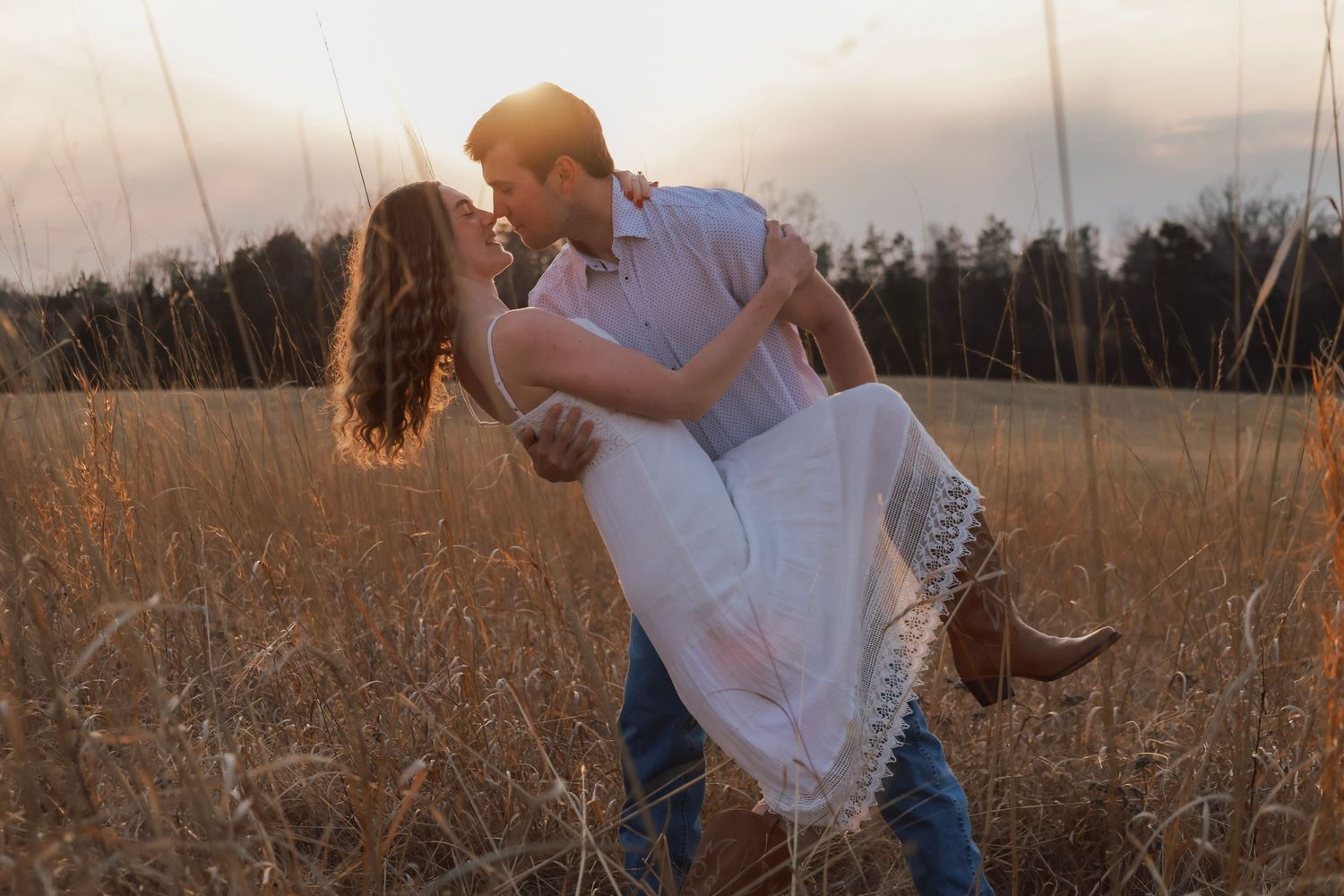 Couple sharing romantic kiss in golden field at sunset, man lifting woman in white dress and cowboy boots.