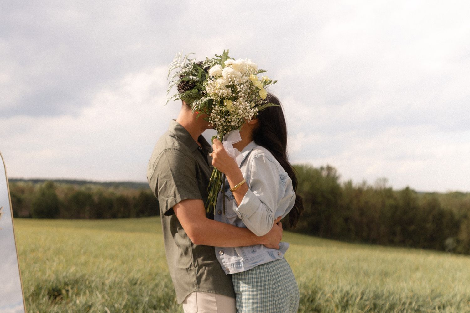 Couple kissing behind a wildflower bouquet in a green field under a cloudy sky.