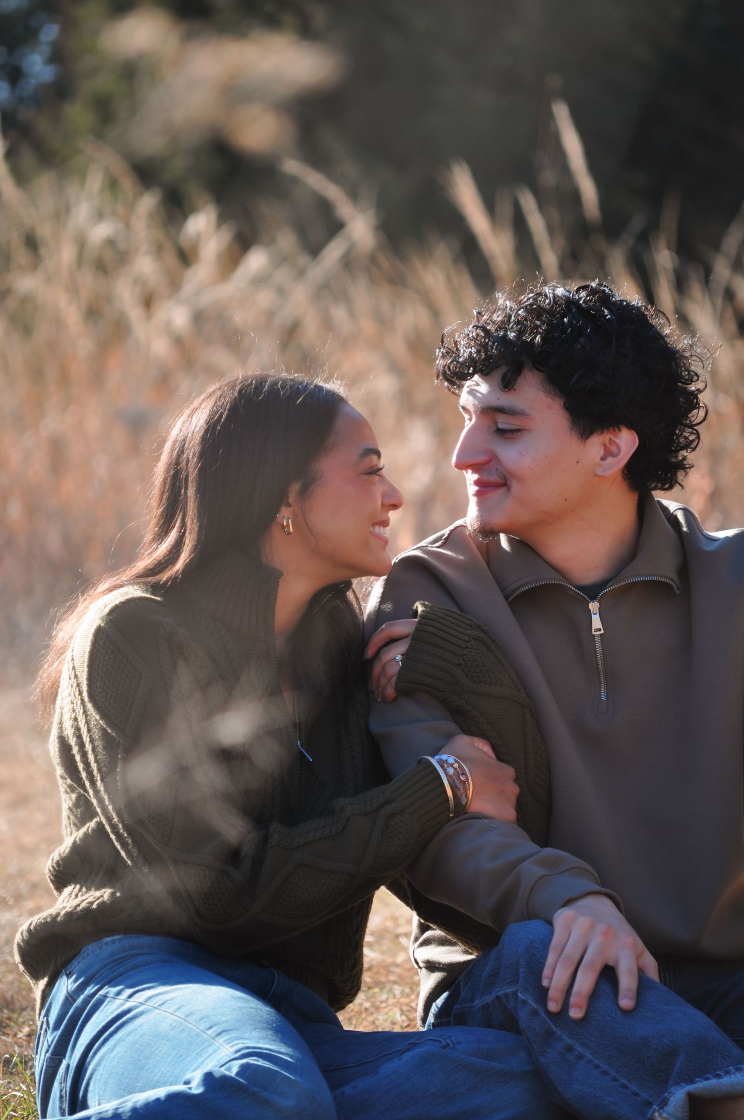 A young couple sitting outdoors in a sunny field, smiling at each other affectionately.