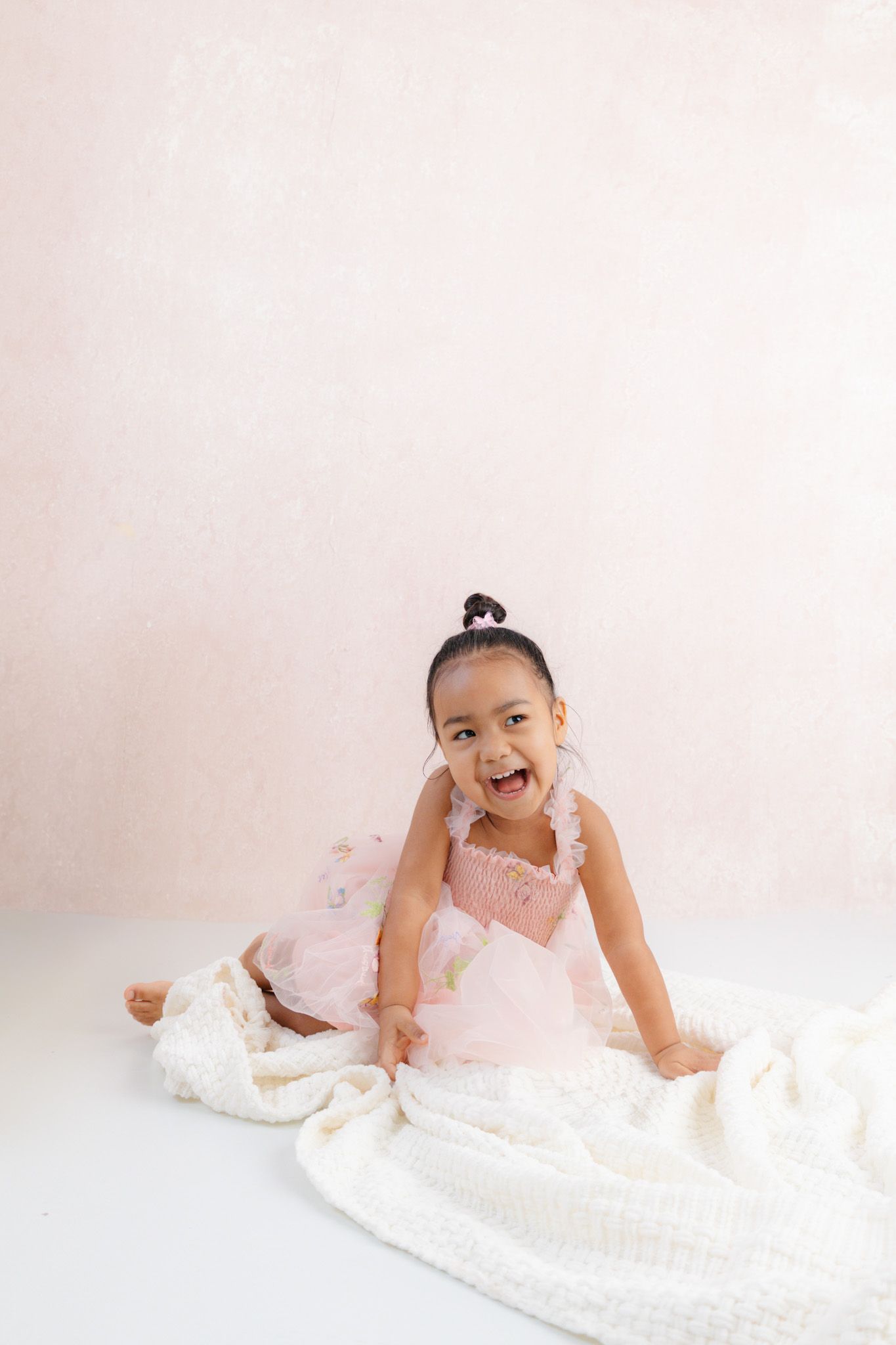 Joyful toddler girl in pink tutu dress sitting on white blanket, laughing, studio portrait with pink background.