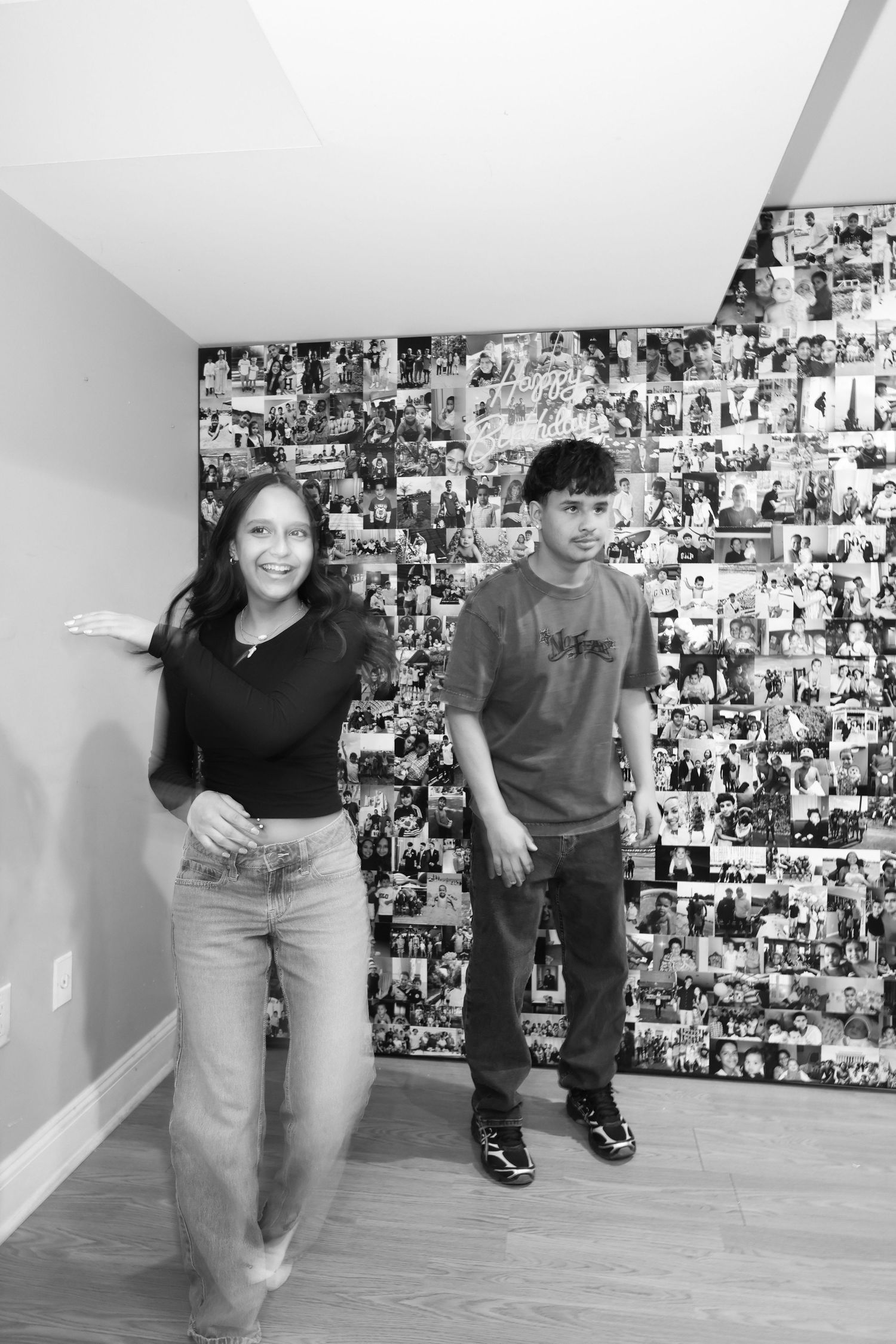 Two siblings smiling in front of a large photo collage wall in a black and white portrait photo.