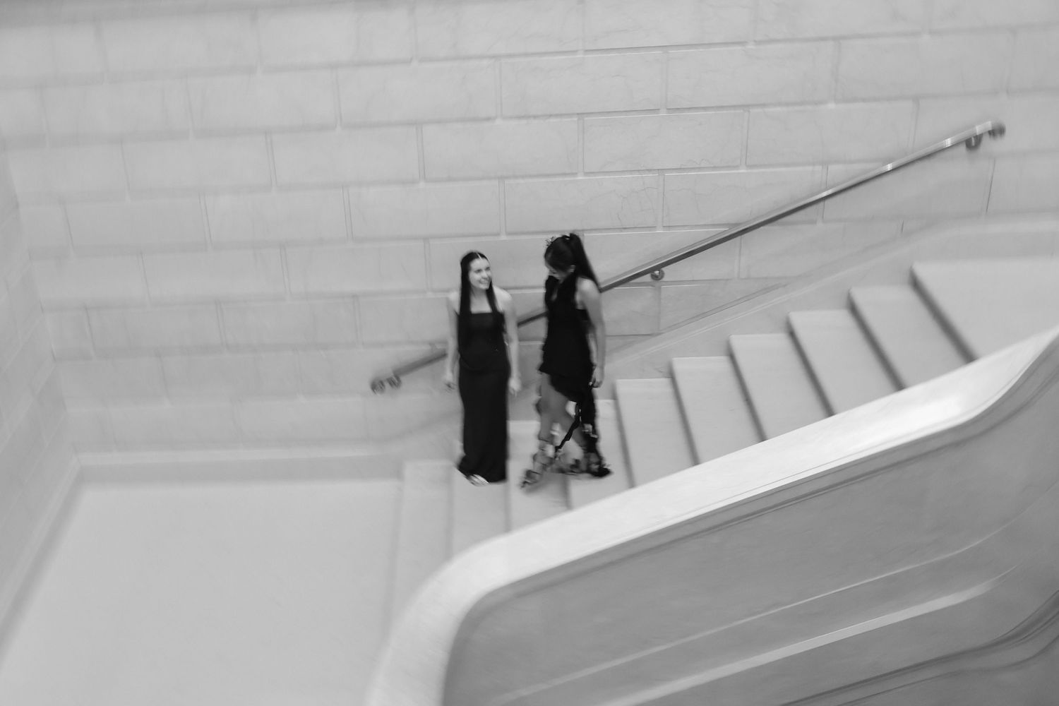 Two women in black dresses converse on white marble stairs in a black and white architectural photo.