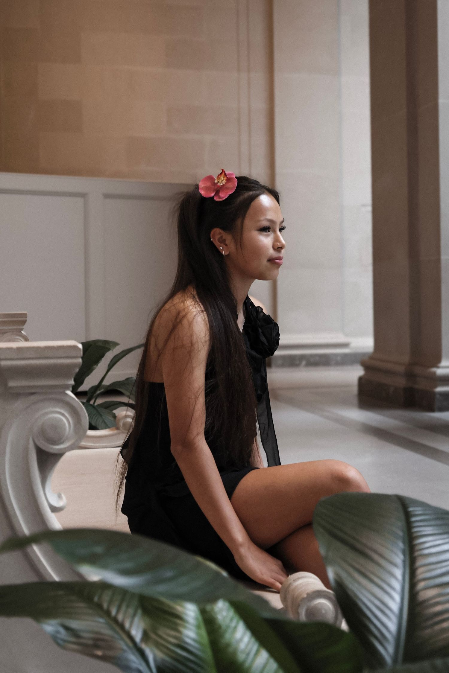 Young woman in black dress with pink flower in hair sits gracefully beside ornate column in elegant interior.