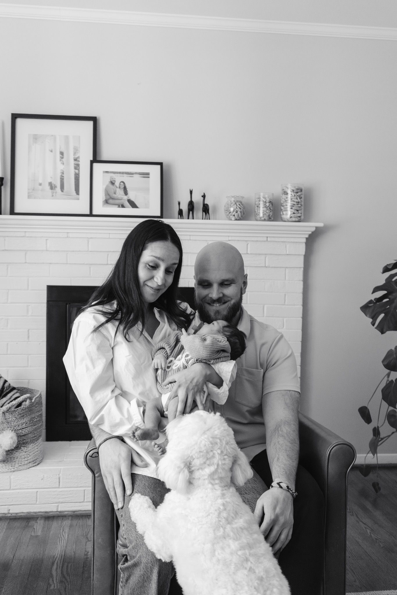 Black and white photo of couple with newborn baby and white poodle dog sitting by brick fireplace at home.