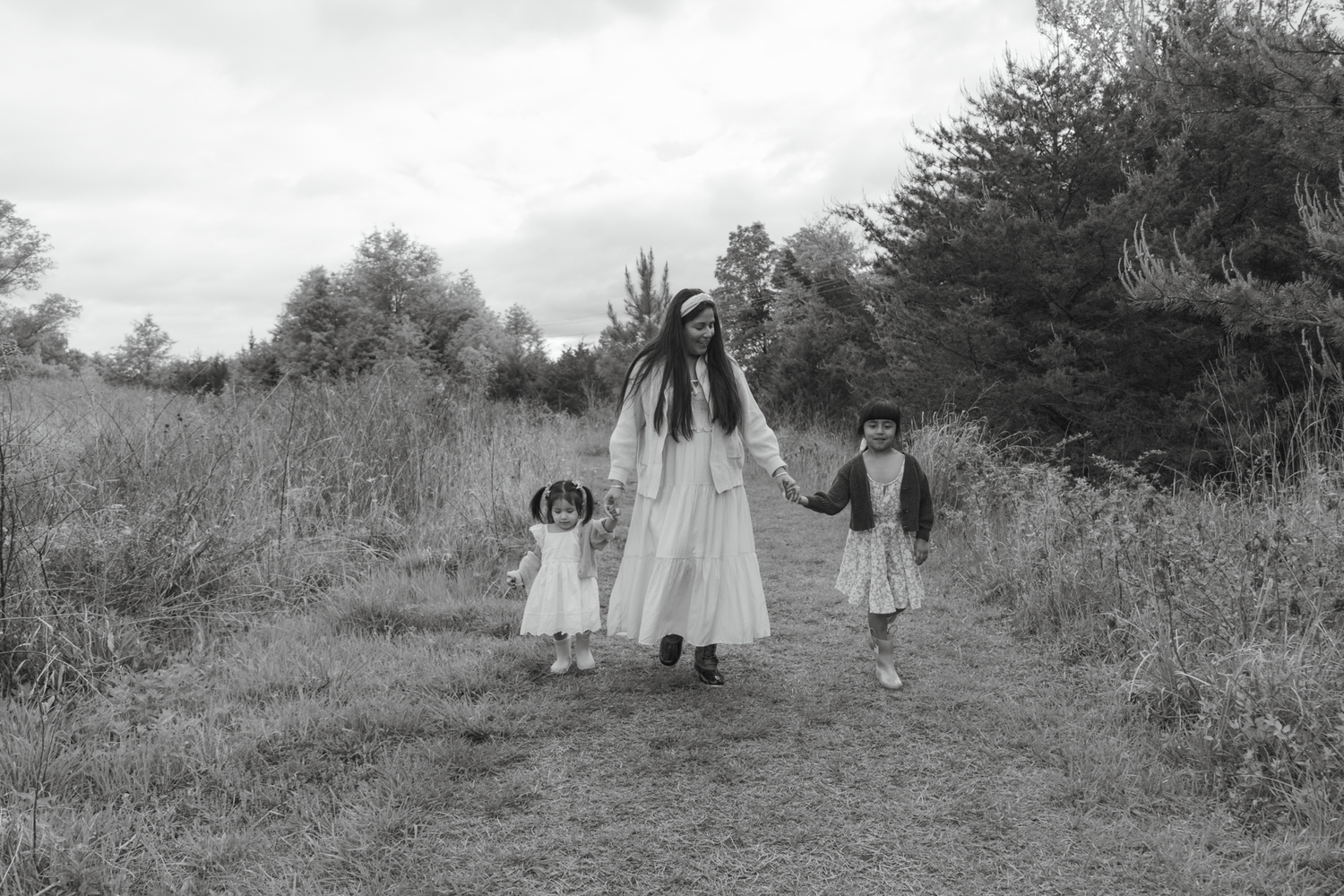 Black and white photo of a woman in white dress walking with two young children on a rural path surrounded by trees.