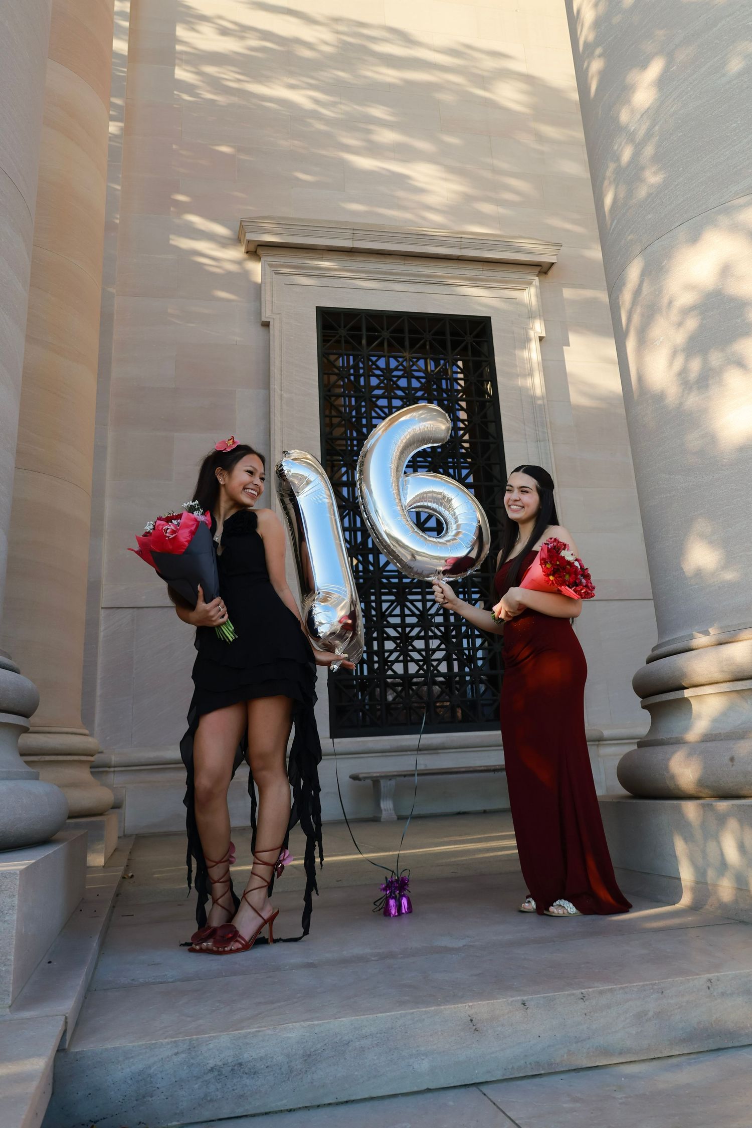 Two young women in formal dresses hold silver number 16 balloons and red roses on stone steps for a sweet sixteen celebration.