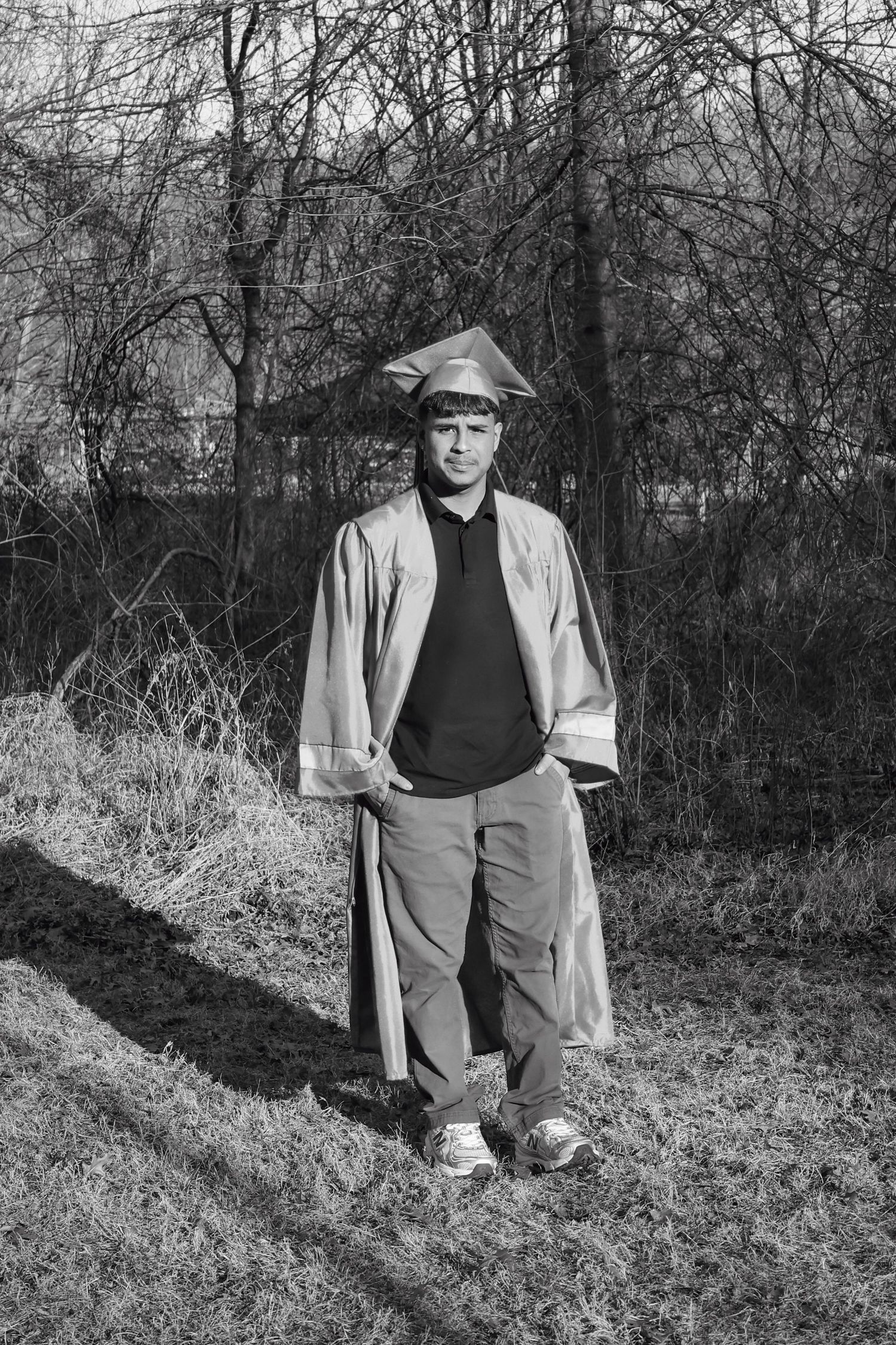 Black and white photo of a young man in graduation cap and gown standing outdoors among bare trees.