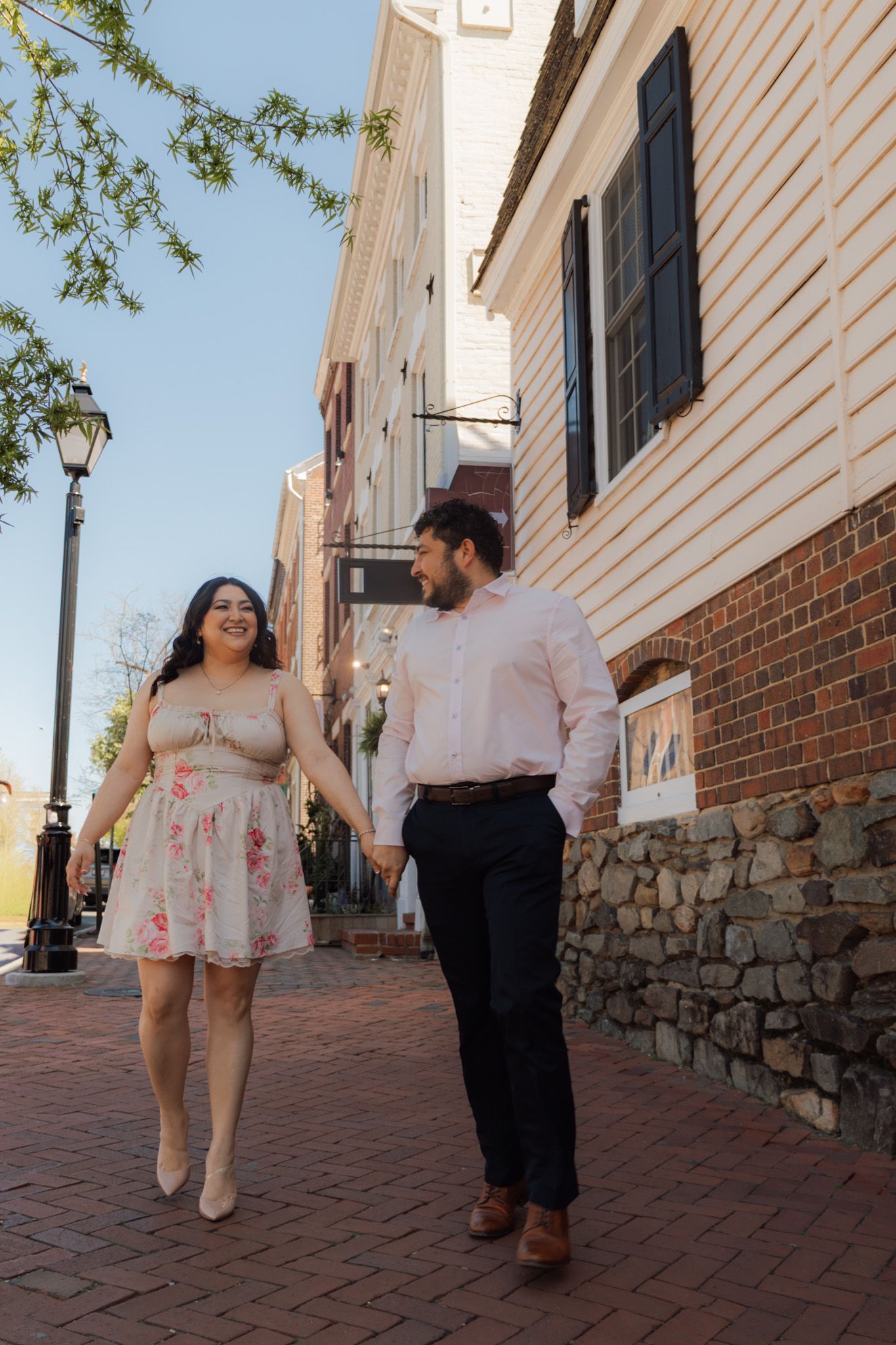 A couple walks hand-in-hand on a brick sidewalk in a historic town, smiling at each other on a sunny day.