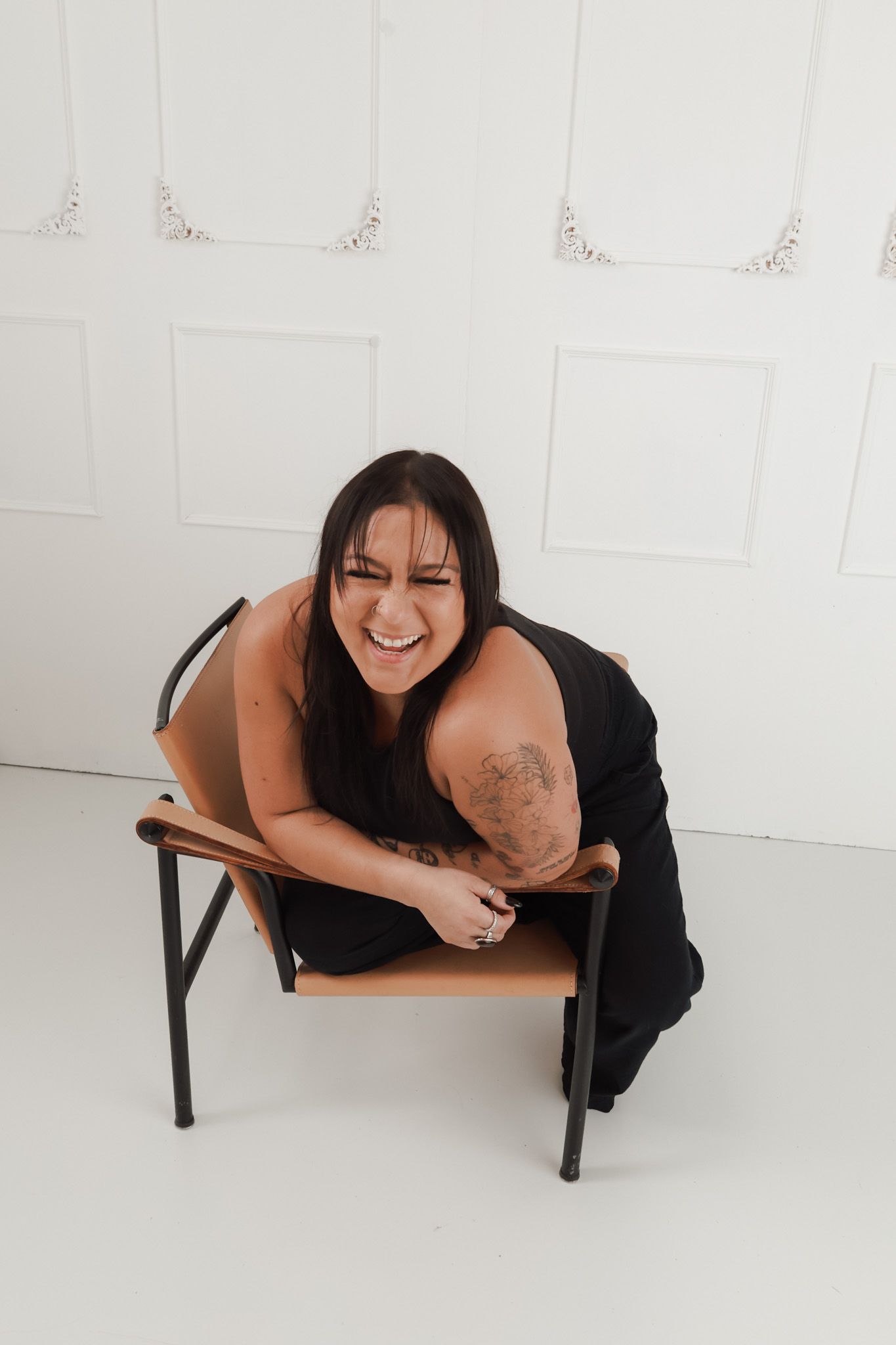 Laughing woman with tattoos in black dress sitting on a tan and black chair against a white wall.