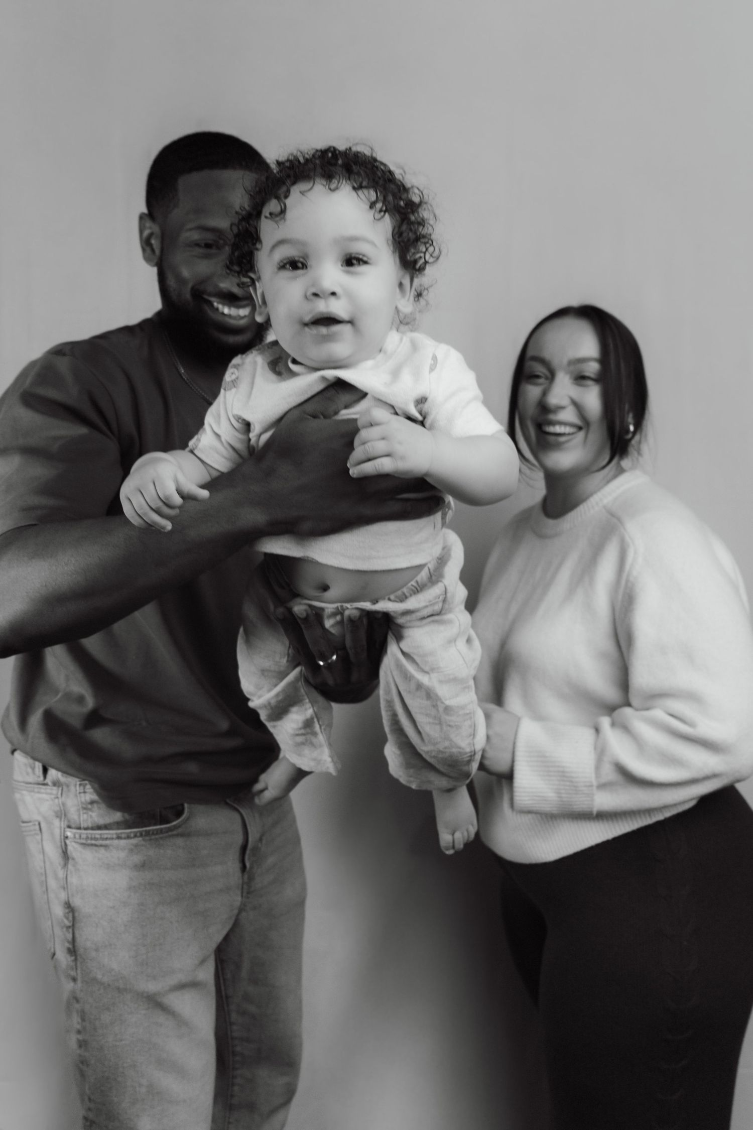 Black and white family portrait of parents joyfully lifting their curly-haired baby against a light background.