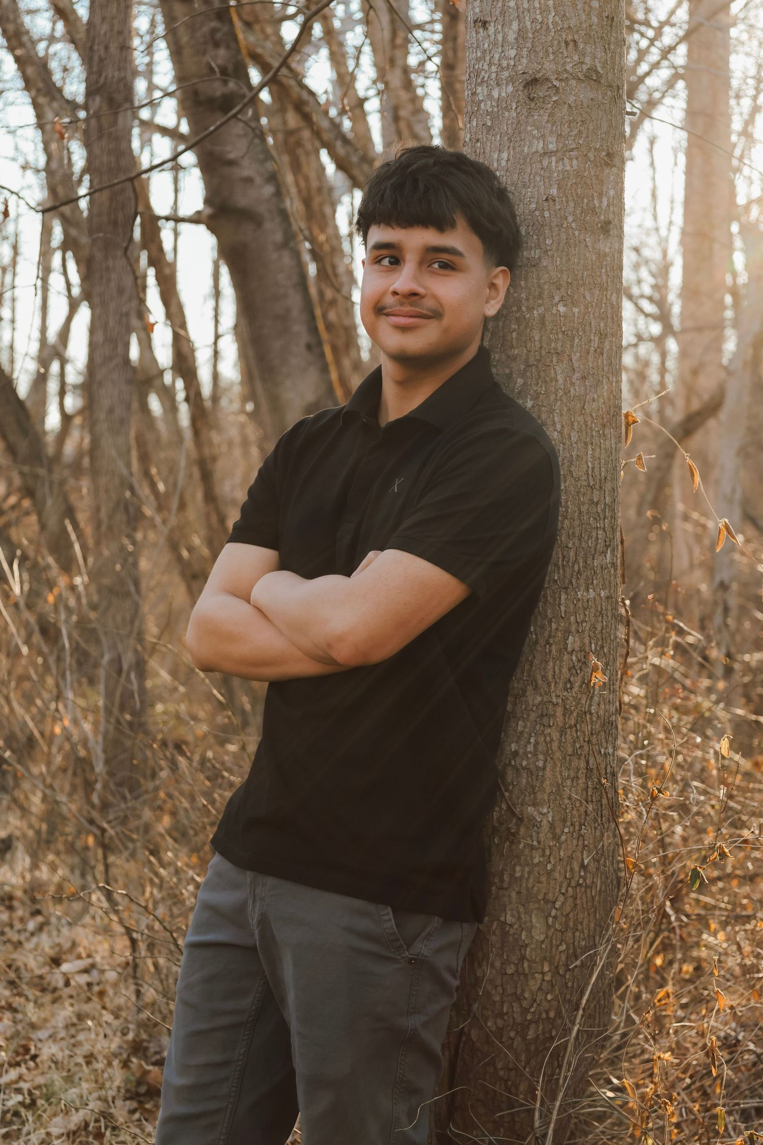 Young man in black polo shirt leaning against a tree with arms crossed in an autumn forest setting.