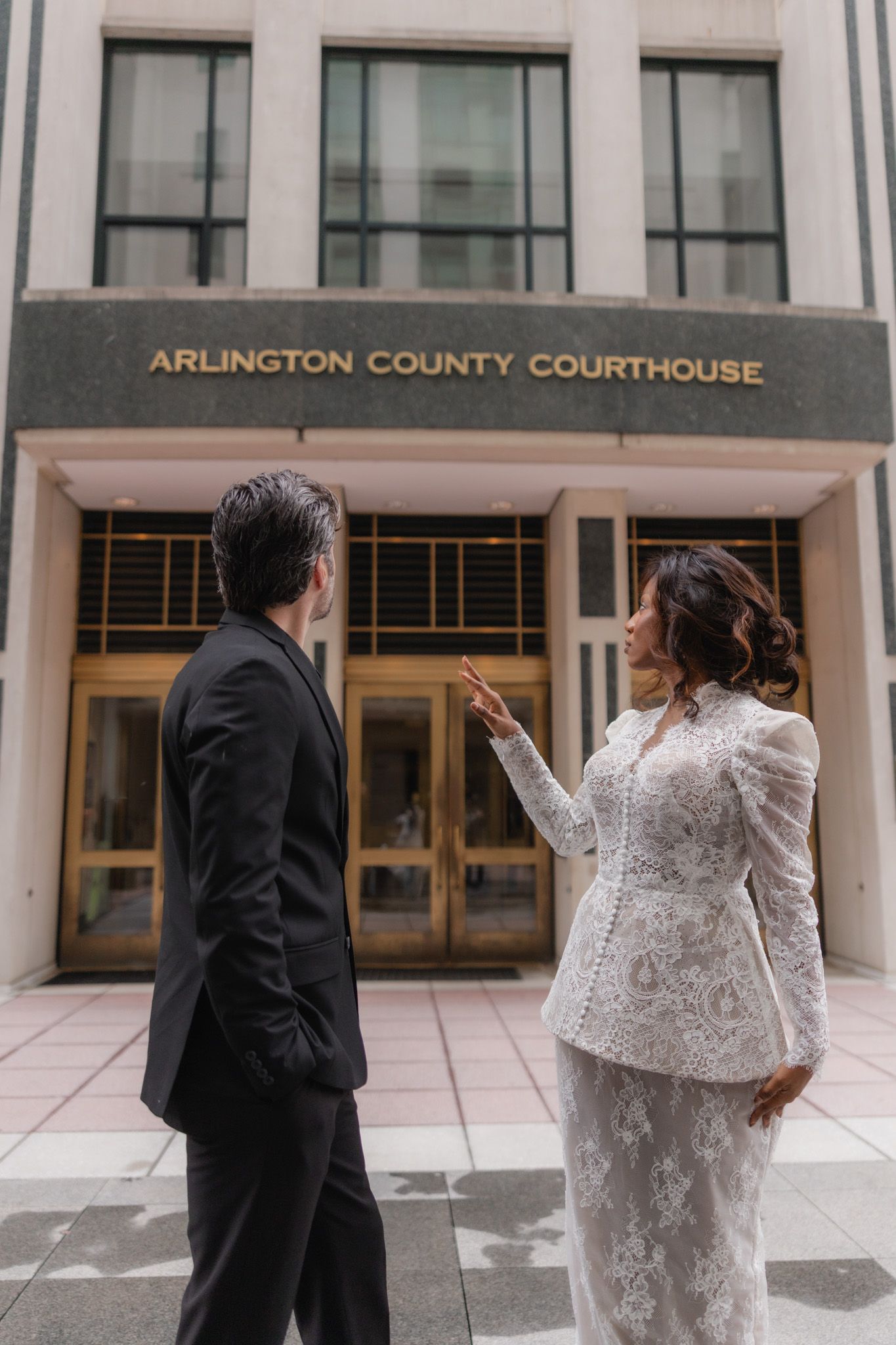 Bride and groom stand before Arlington County Courthouse in wedding attire, bride pointing at the building entrance.