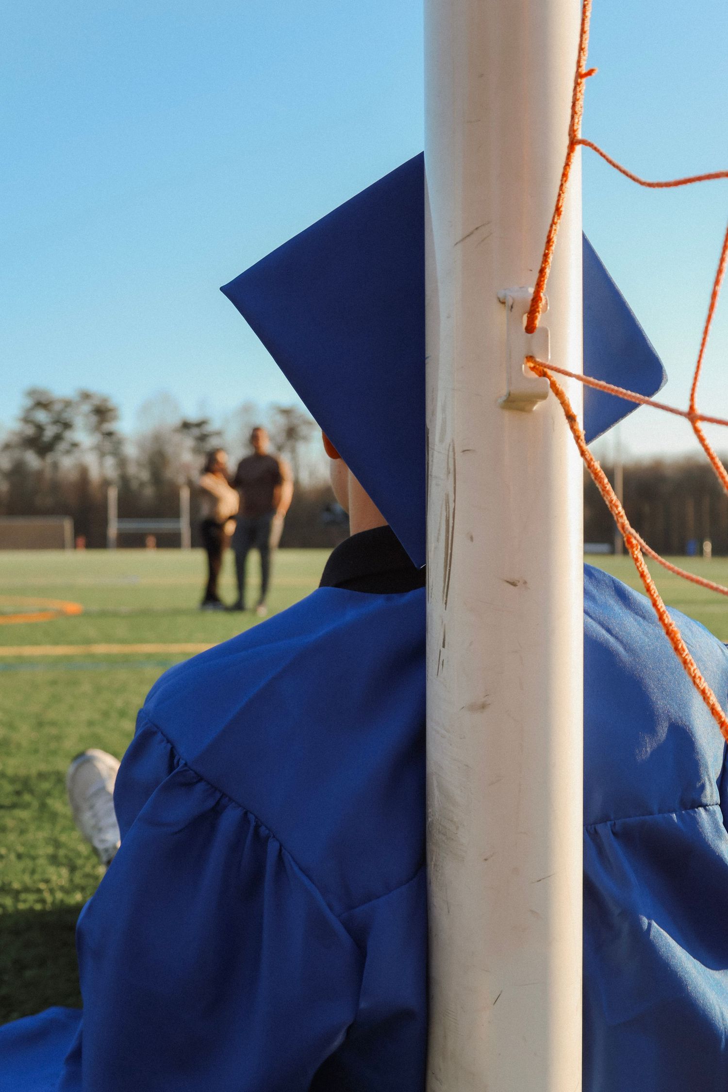 Graduate in blue cap and gown stands by soccer goal post on green field under clear blue sky. parents into the distance