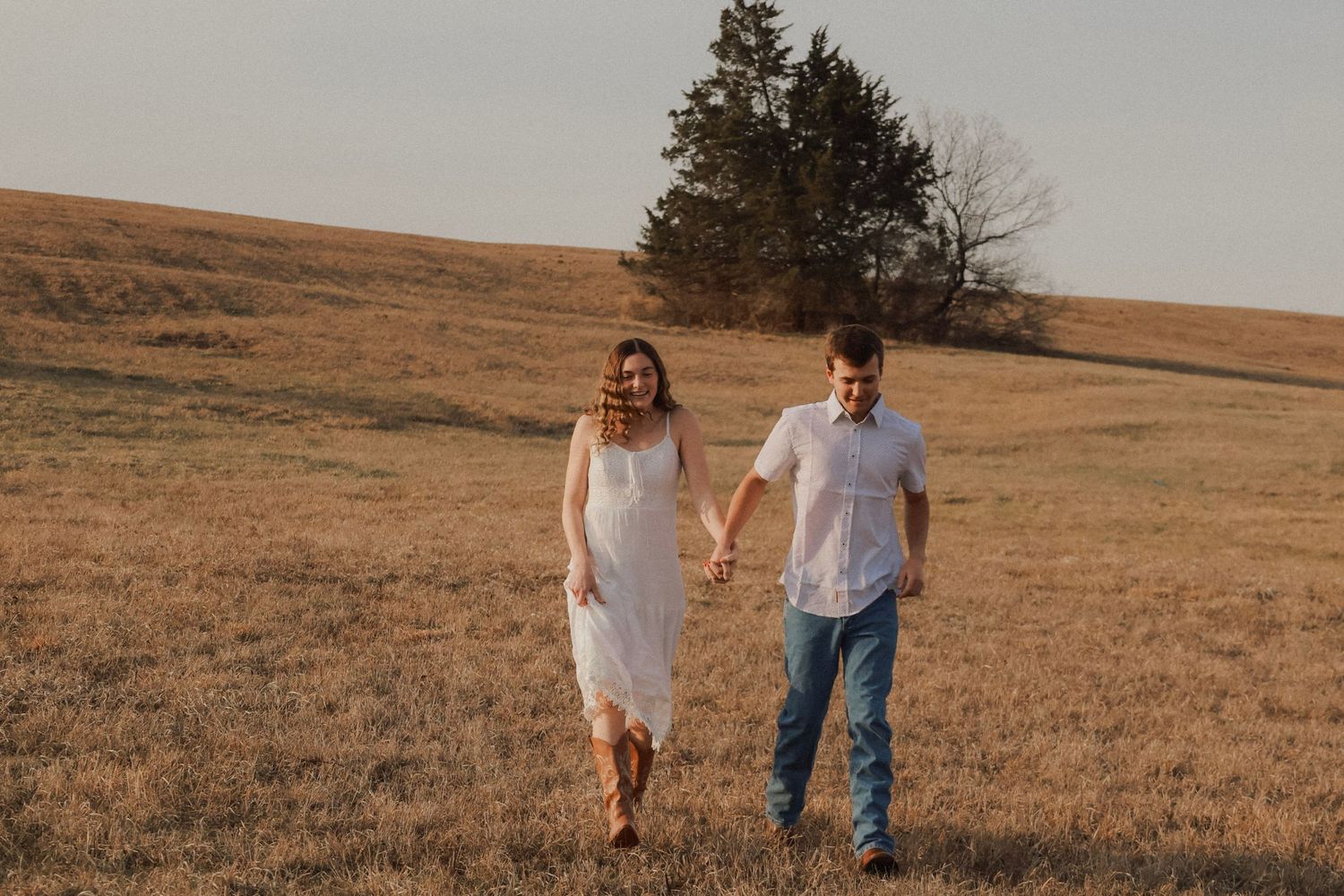 A couple holds hands and walks through a golden field at sunset, woman in white dress and man in blue jeans.