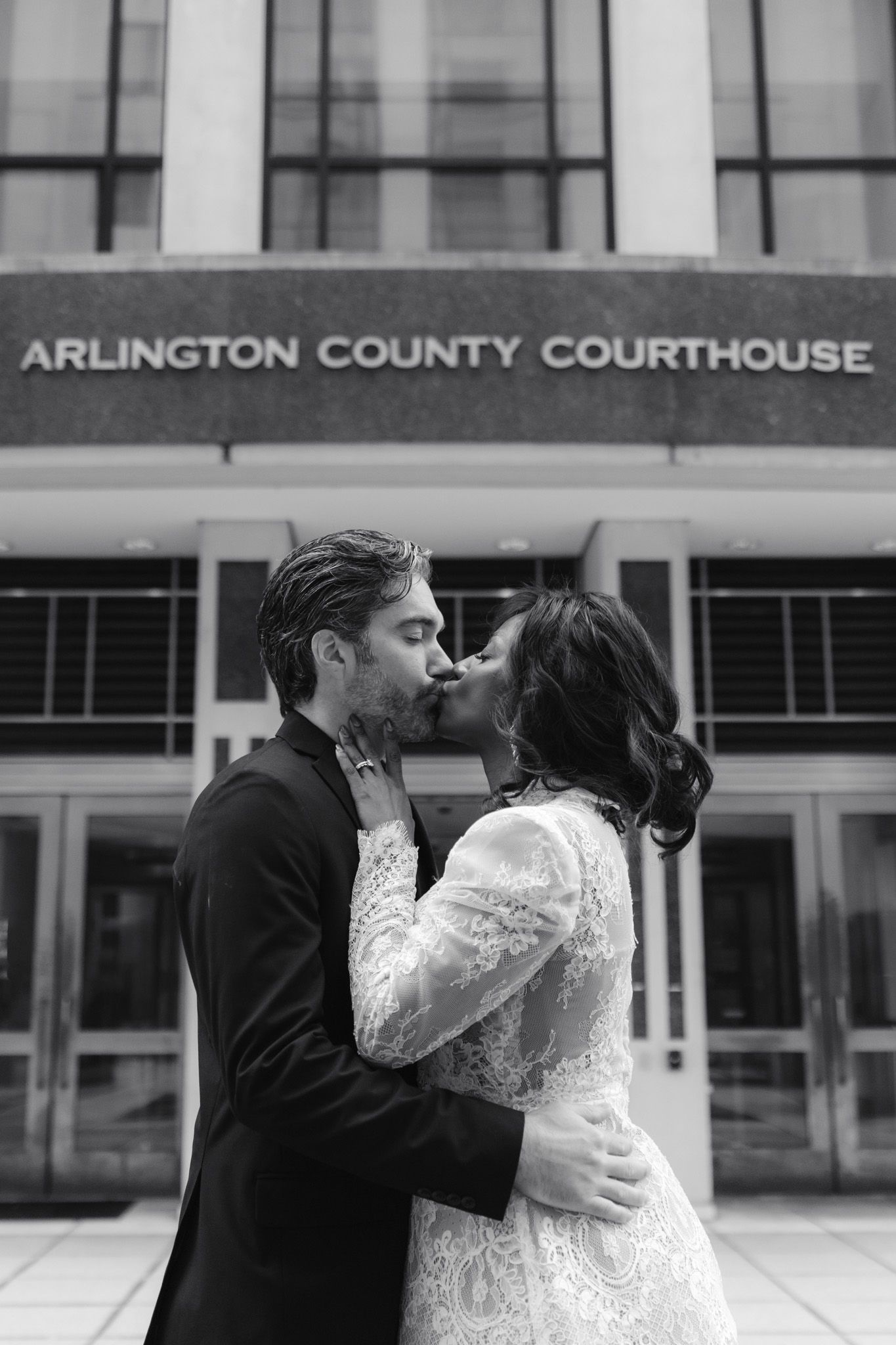 Couple kissing in wedding attire outside Arlington County Courthouse in black and white photo.