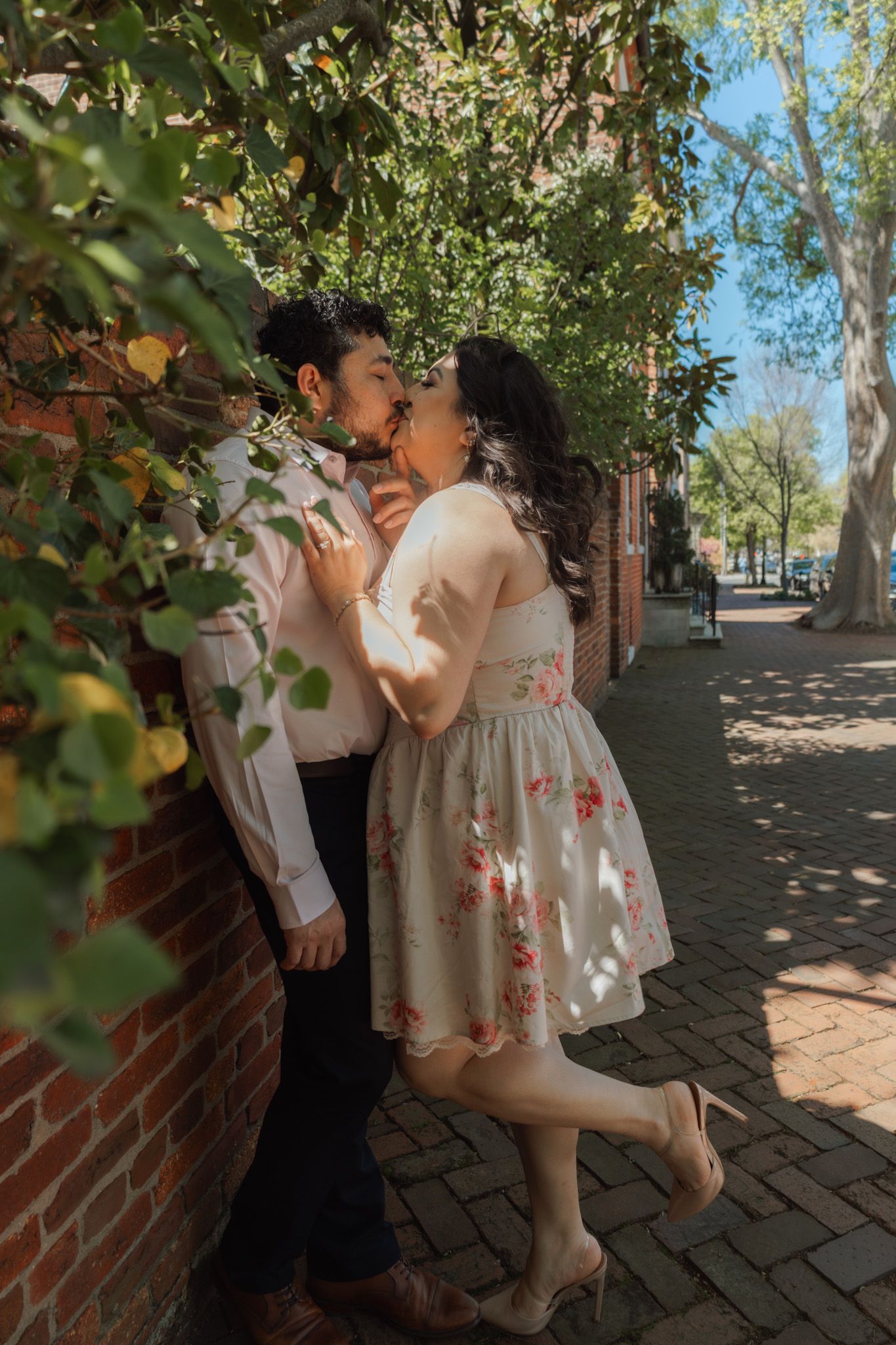 Couple sharing a romantic kiss against a brick wall on a sunny tree-lined brick sidewalk, woman in floral dress.
