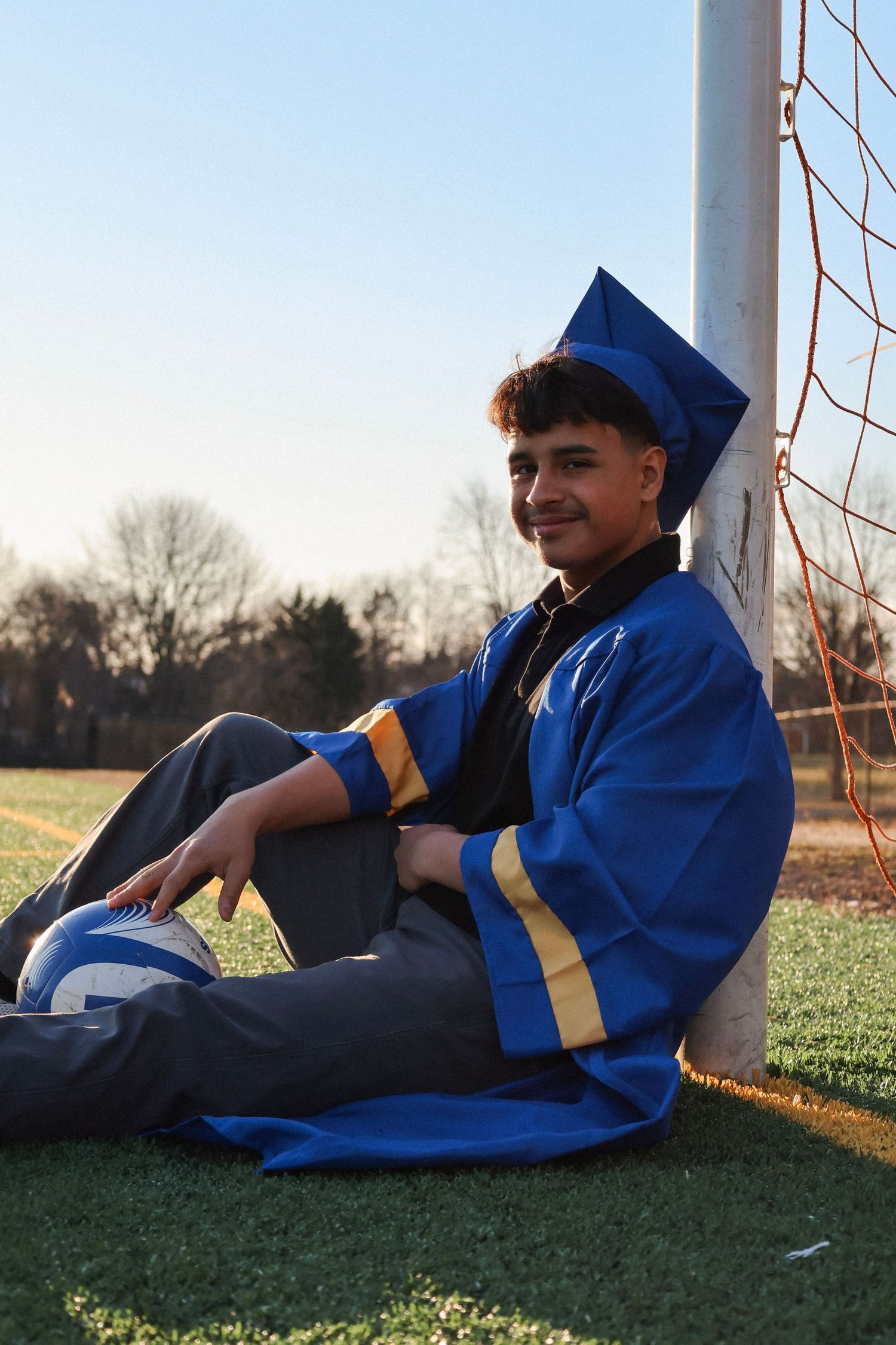 Young man in blue graduation cap and gown sitting by a soccer goal post on a green field, smiling.