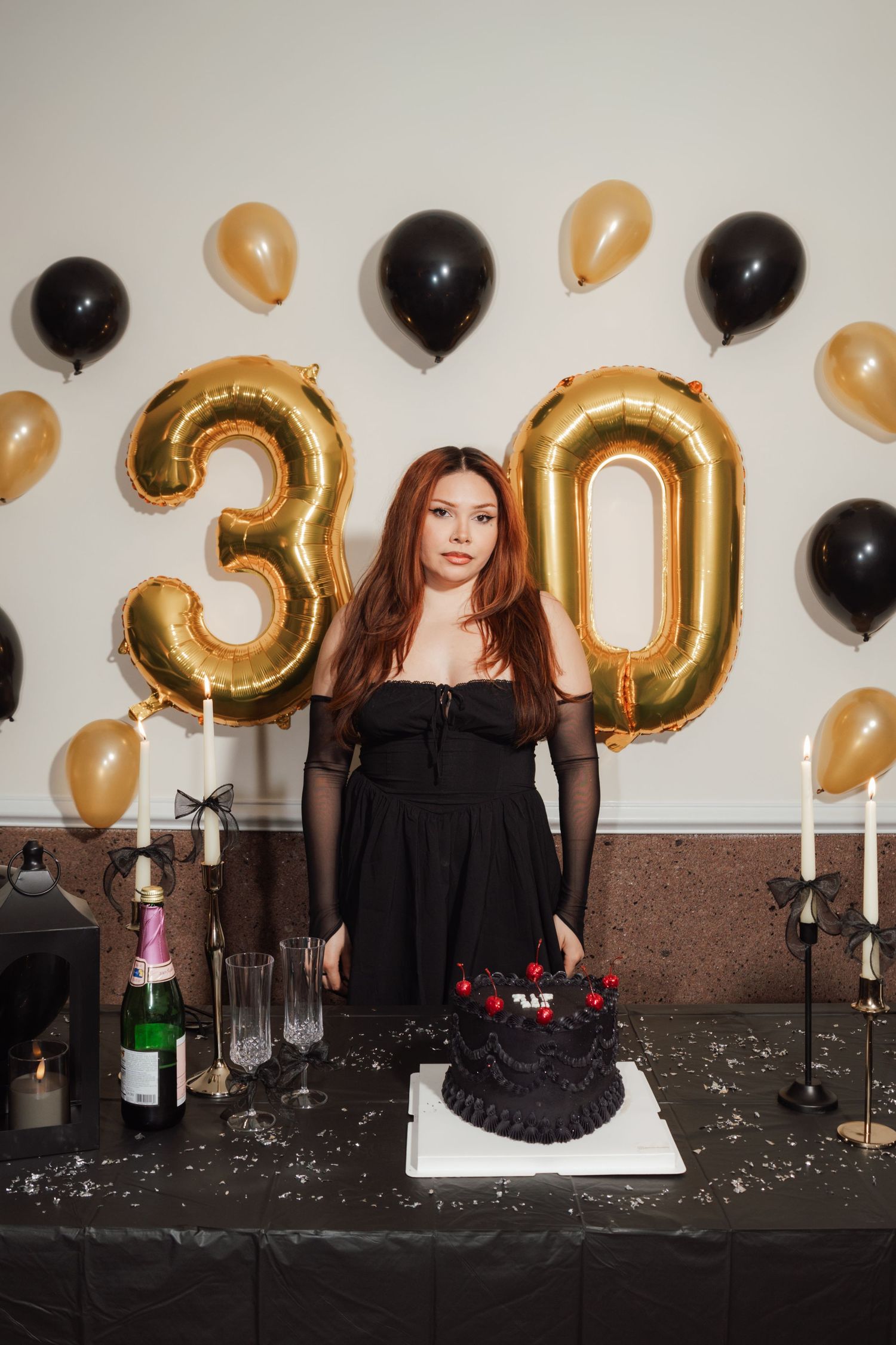 Woman in black dress celebrating 30th birthday with black cake, champagne, candles, and gold and black balloons.
