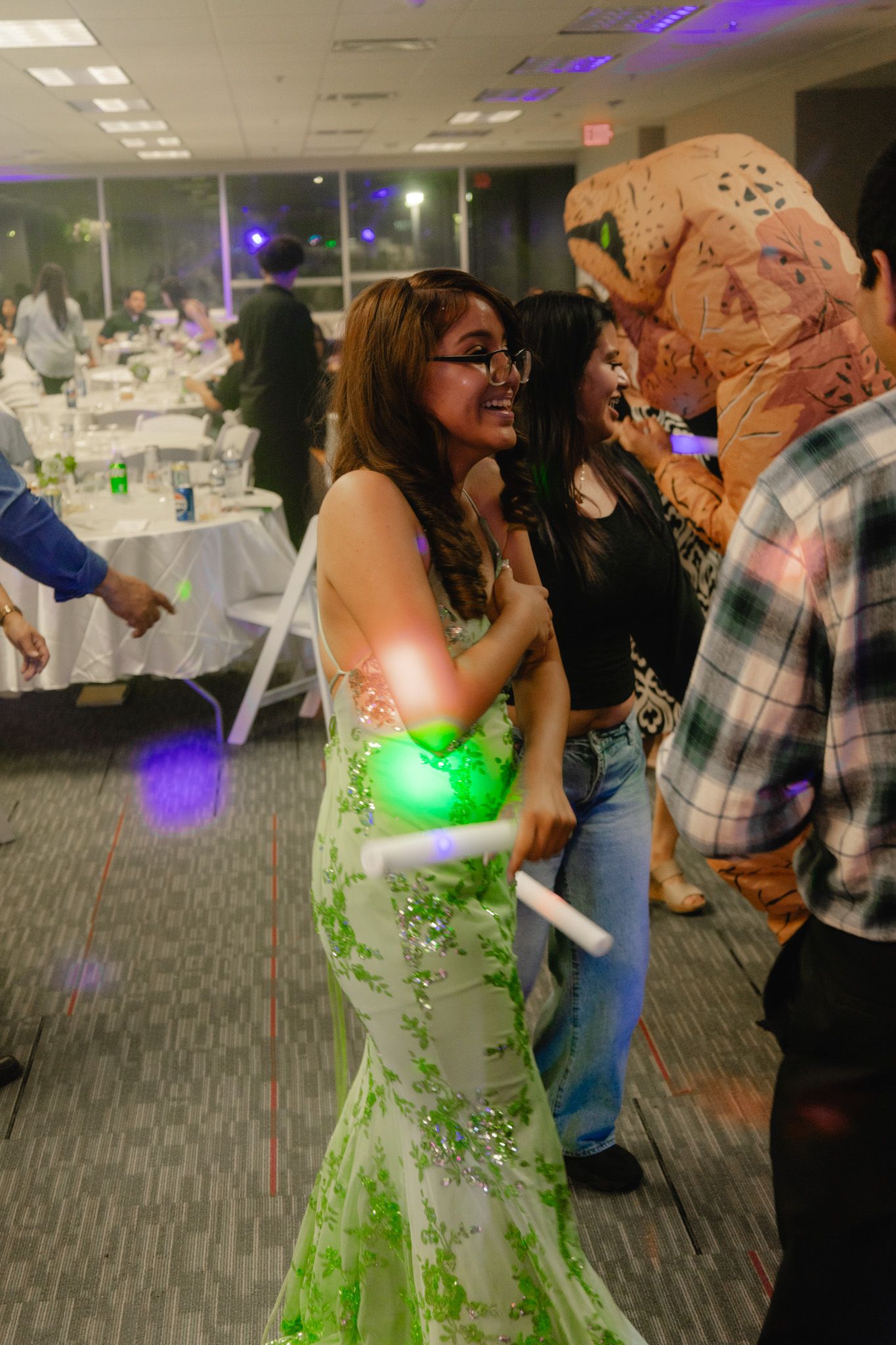 Woman in green floral gown dancing with glow sticks at an indoor party event with guests and decorations.