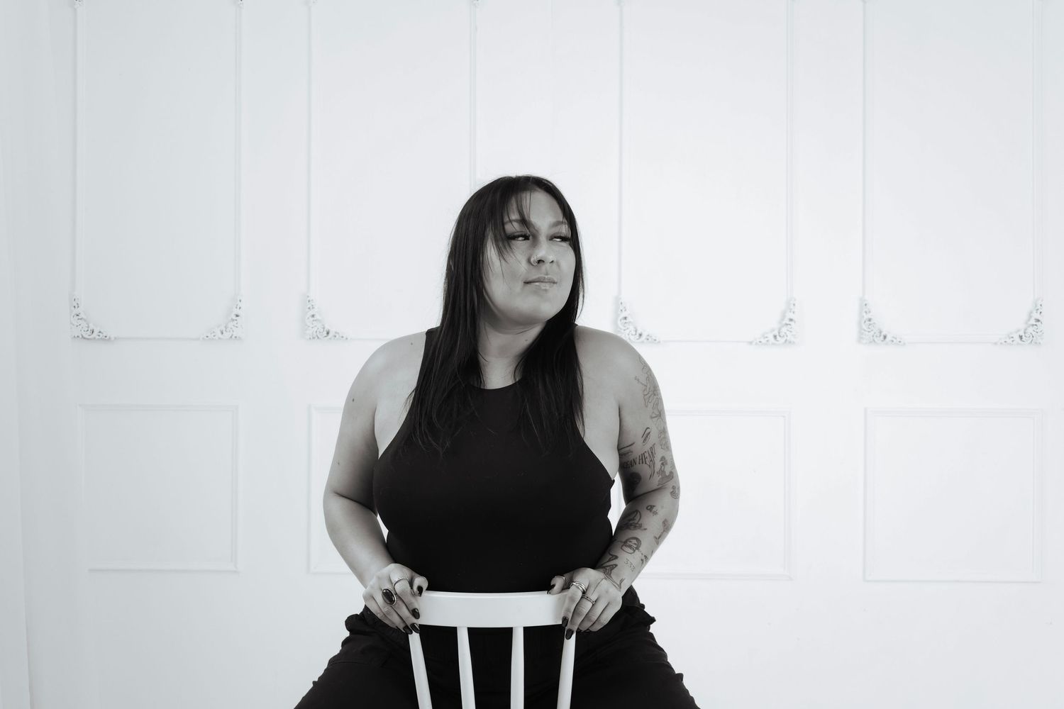 Black and white portrait of a woman with long dark hair sitting on a white chair against a white wall.