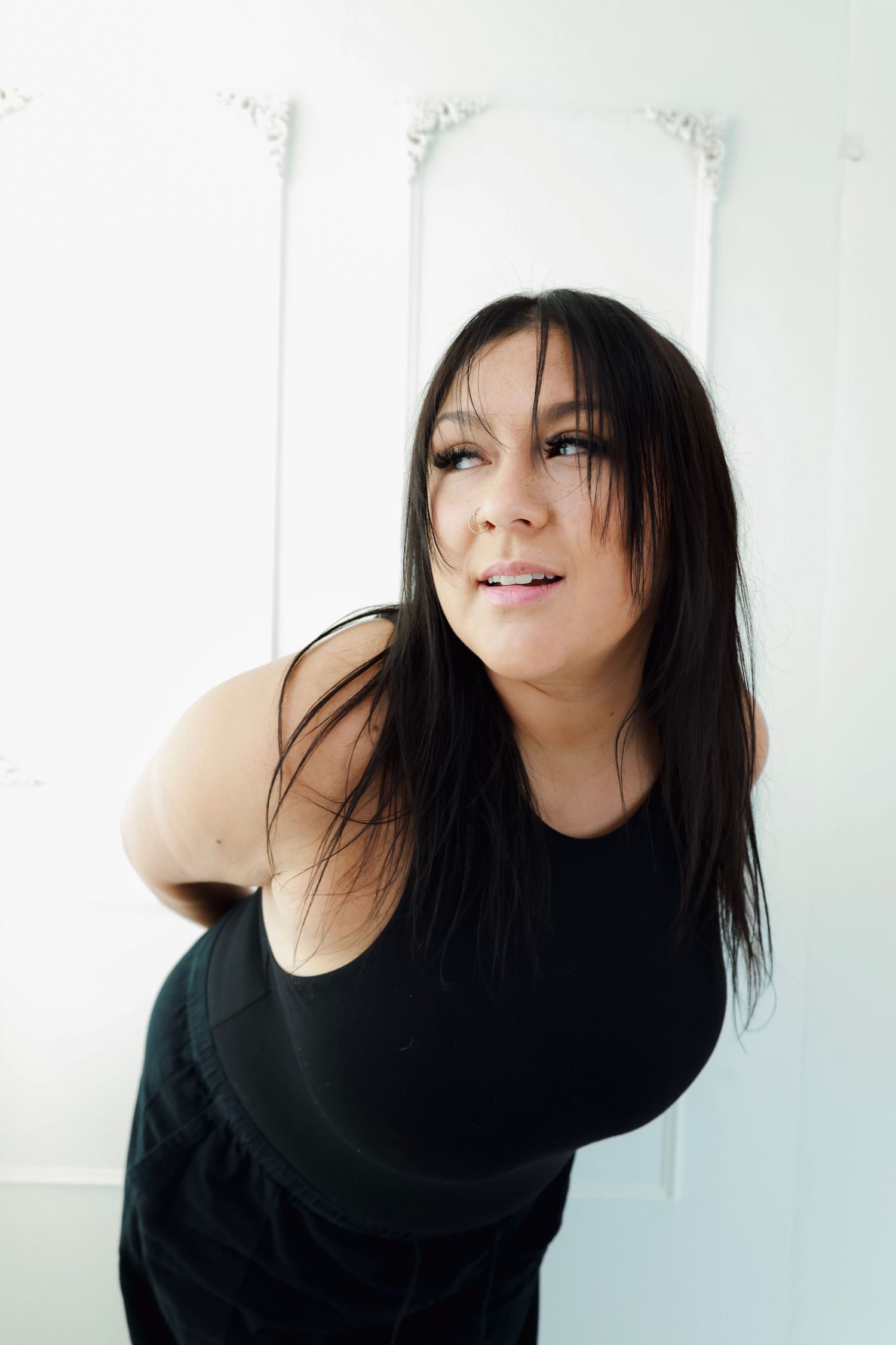 A young woman with long dark hair wearing a black sleeveless top leans forward against a white decorative wall in a studio.