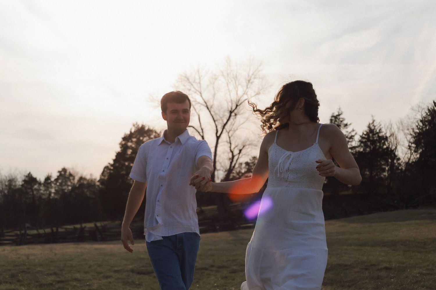 Couple running together in a sunlit field at dusk, woman in white dress and man in white shirt, trees in background.