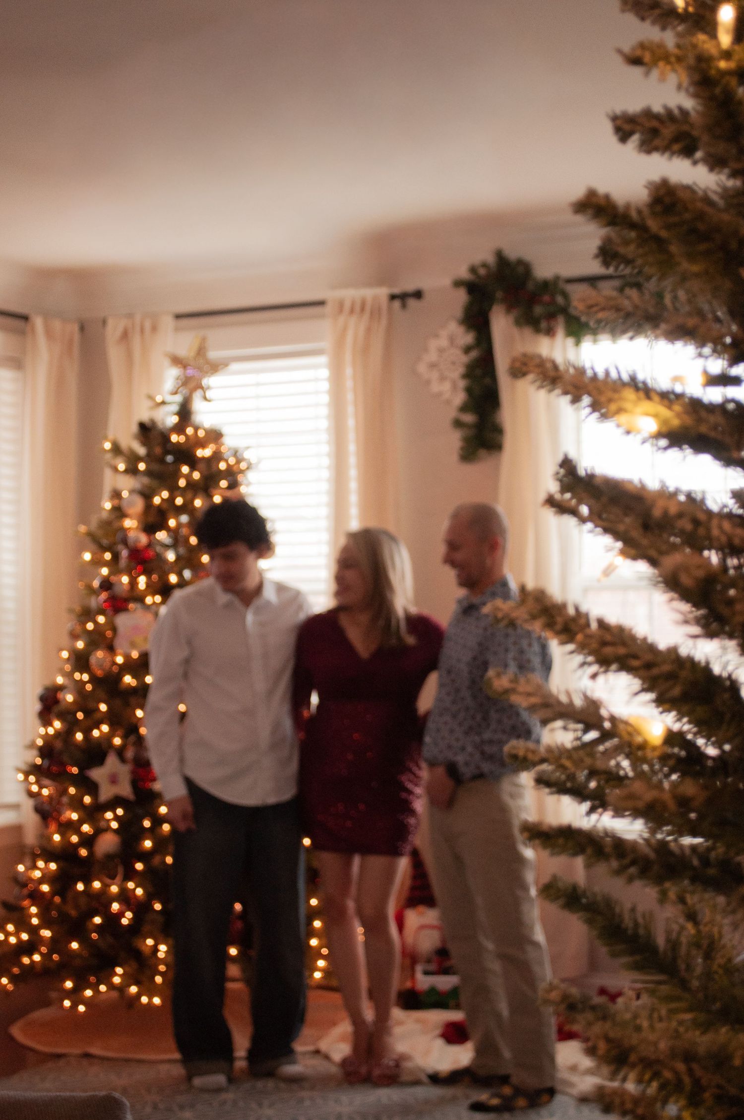 Family of three posing together indoors near decorated Christmas trees with warm twinkling lights during holiday season.