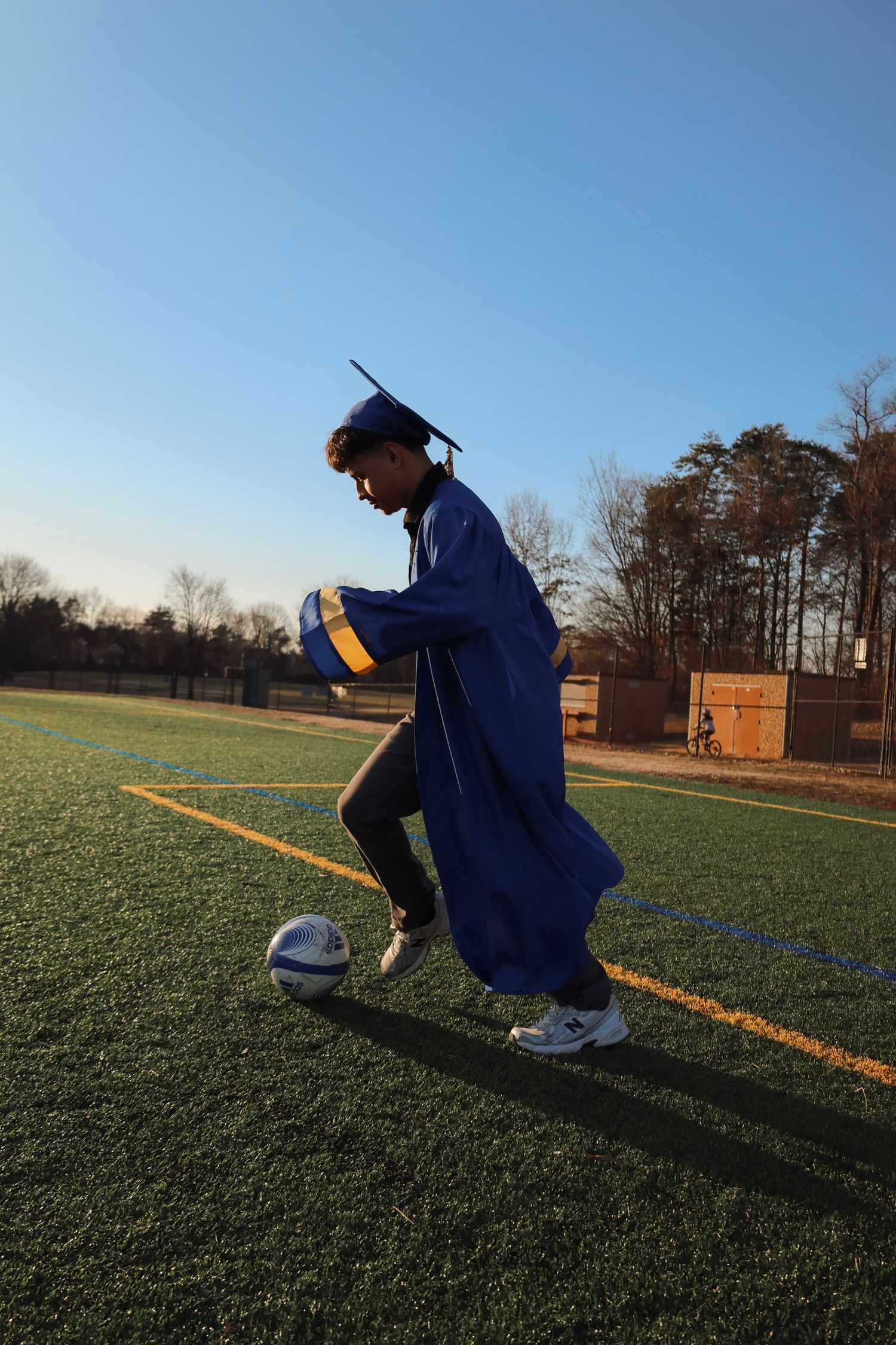 Graduate in blue cap and gown kicking a soccer ball on a turf field under a clear blue sky.