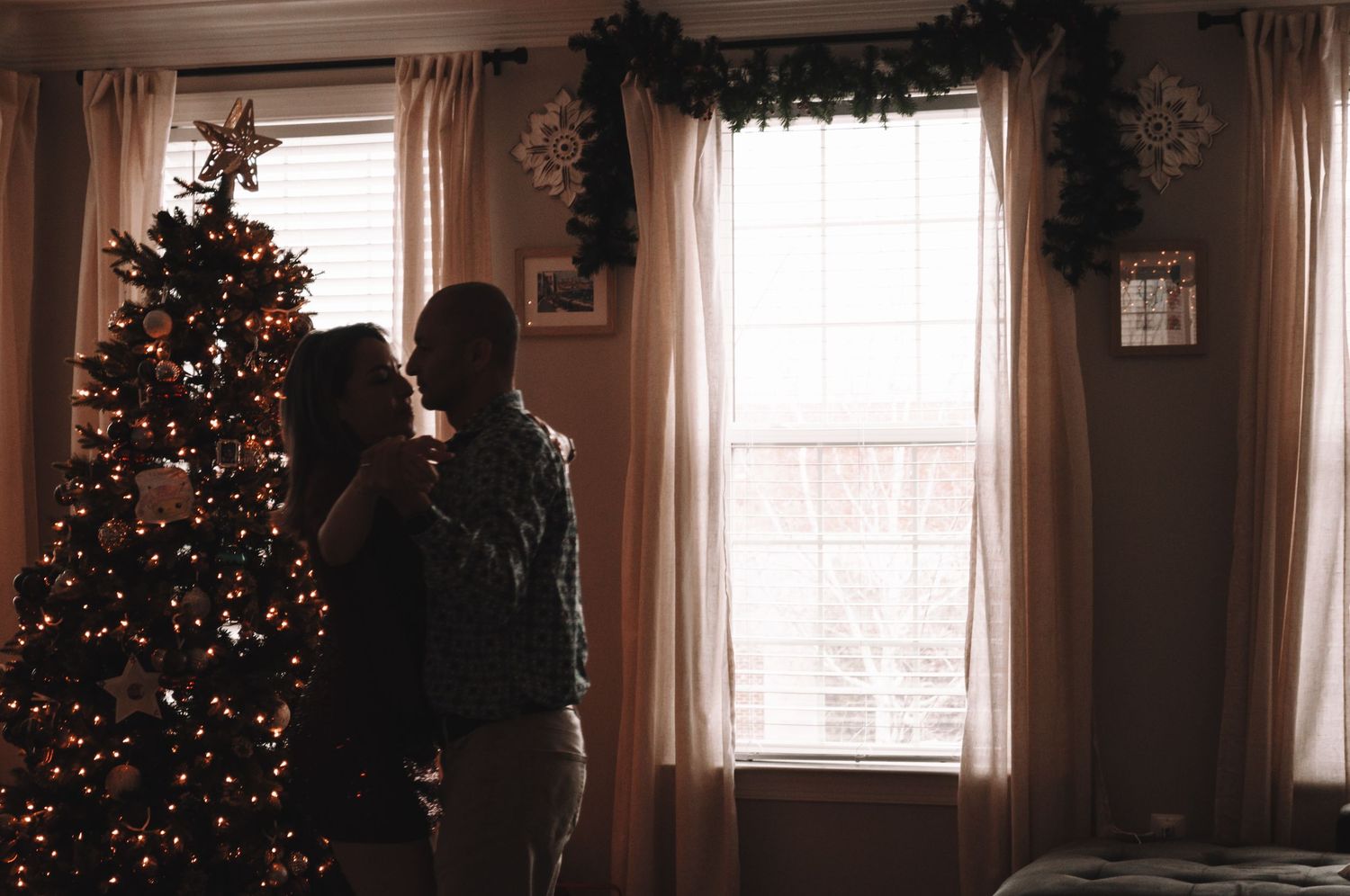Two silhouetted figures stand near a decorated Christmas tree with lights in a cozy holiday home setting.
