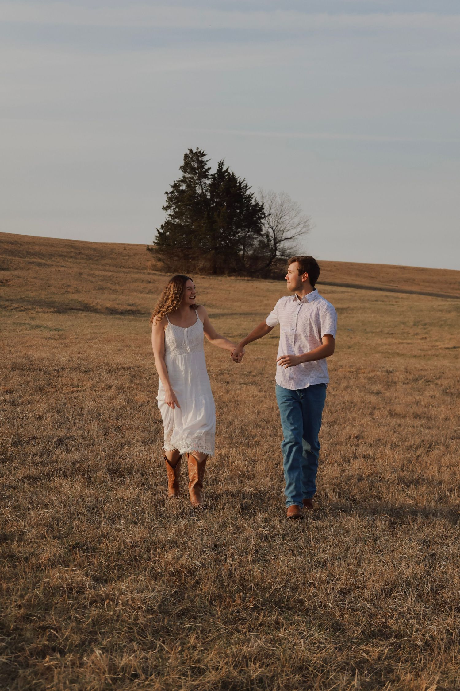 A couple holds hands walking through a golden field at sunset, woman in white dress, man in white shirt and jeans.