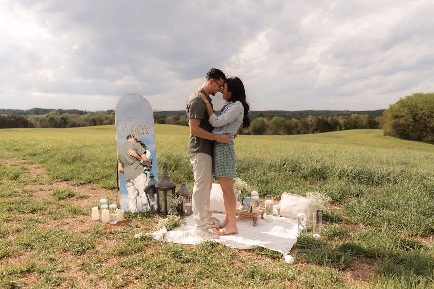 Couple kissing in a green field with romantic picnic setup, candles, and floral arrangements under cloudy sky.