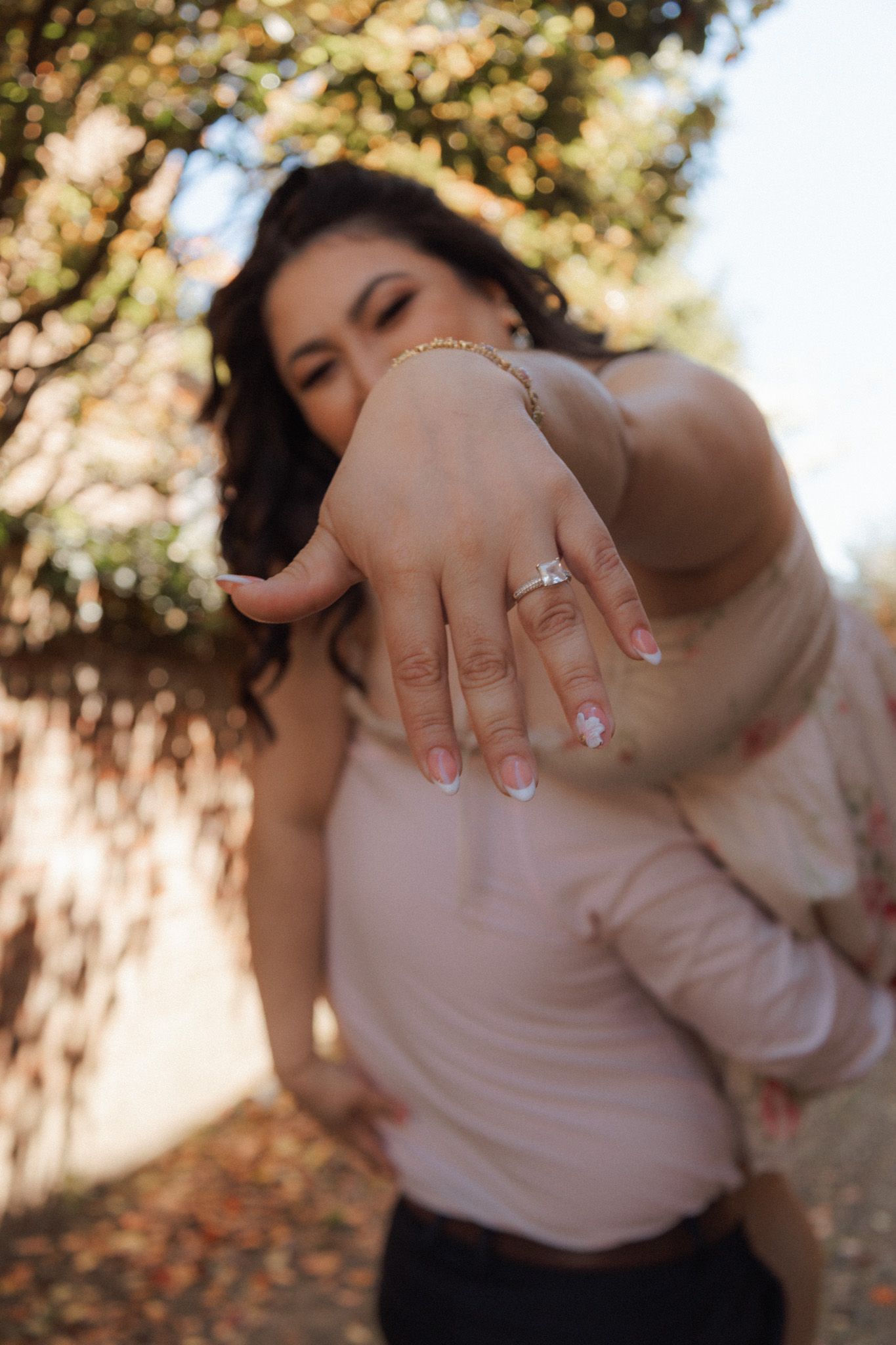 Woman proudly displaying a diamond engagement ring outdoors, with bokeh background of sunlit trees.
