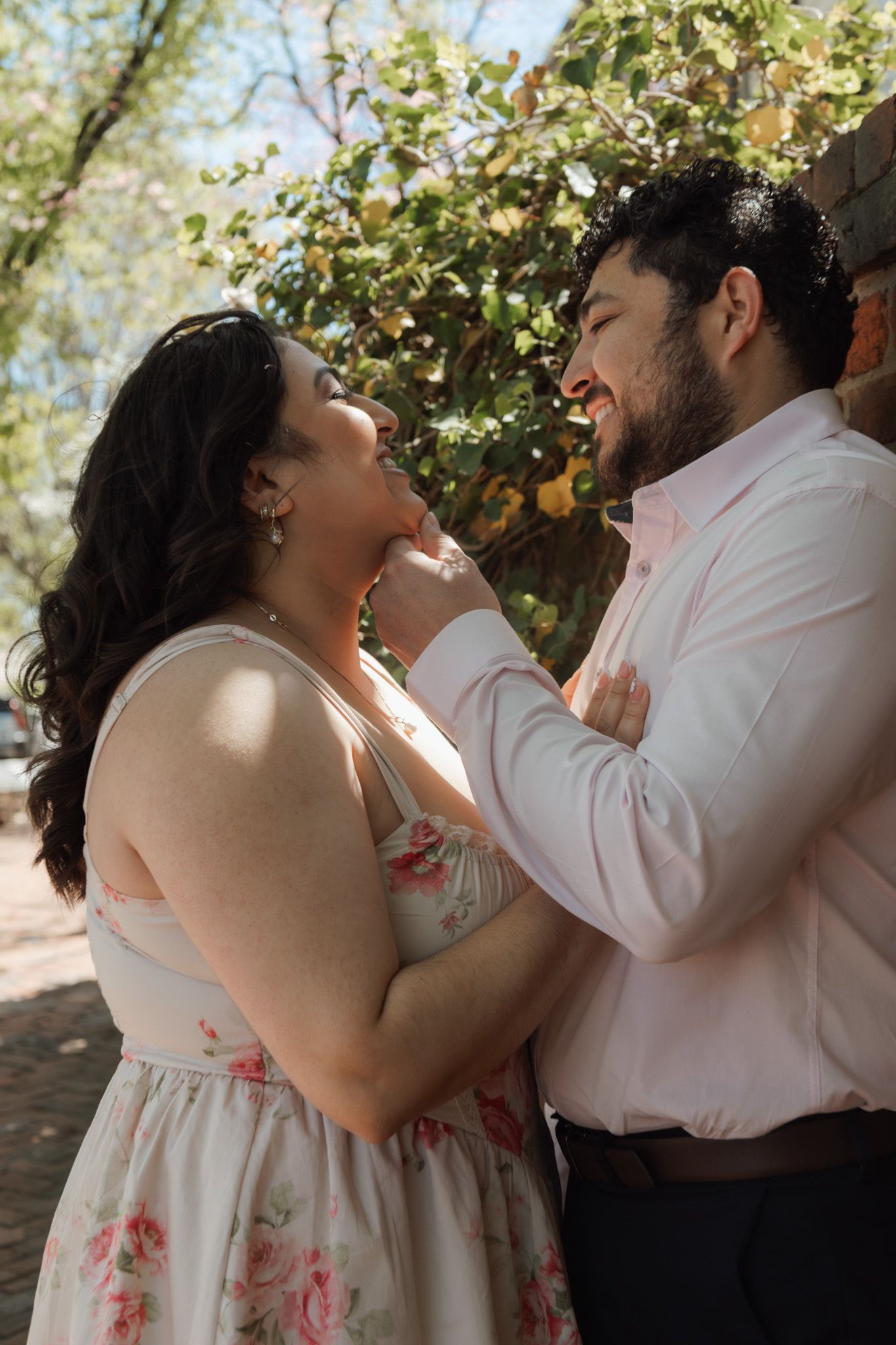 A couple shares a romantic moment outdoors, man in pink shirt gently lifting woman's chin, surrounded by lush greenery.