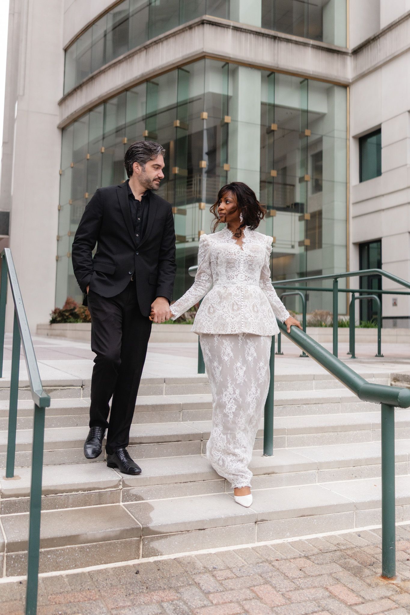 Couple in formal attire holding hands on outdoor steps, man in black suit, woman in white lace two-piece outfit outside courthouse elopement