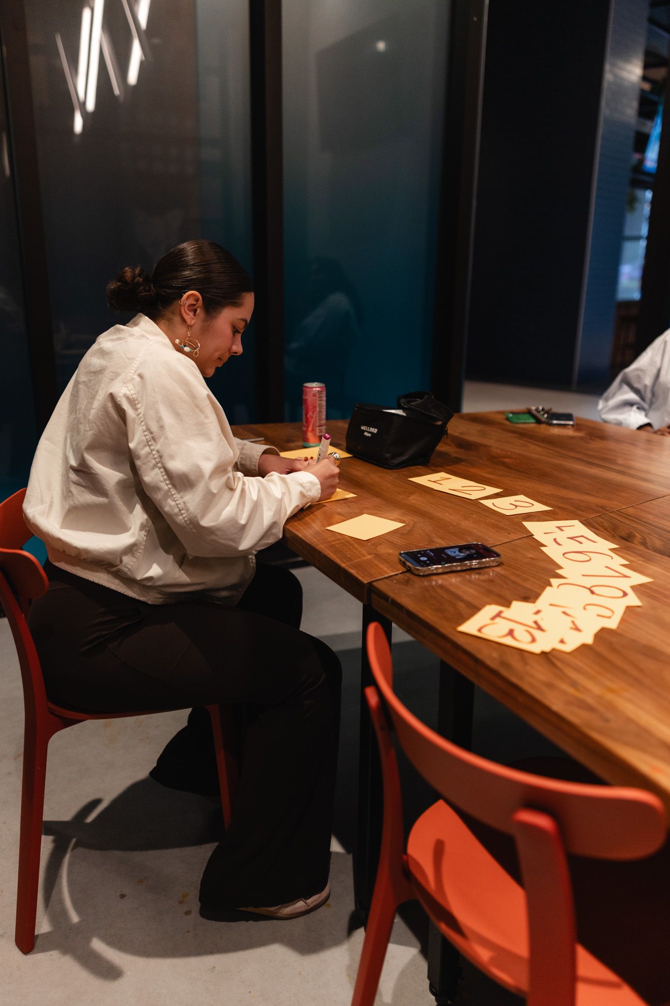 Woman in white shirt writing notes at wooden table with cards and phone in modern office setting.