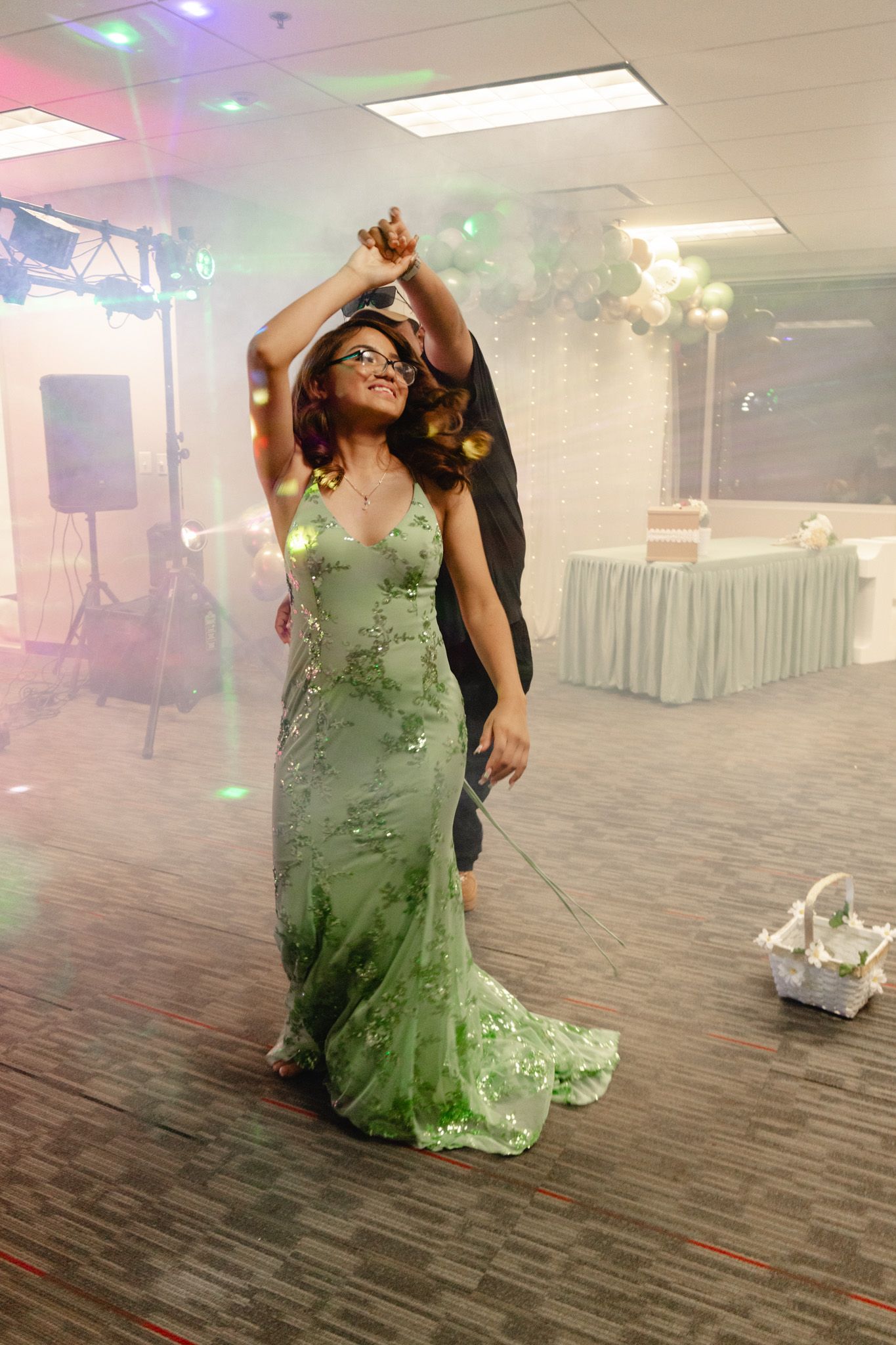 Woman in green sequin gown dancing at formal event with colorful disco lights and balloon arch backdrop.