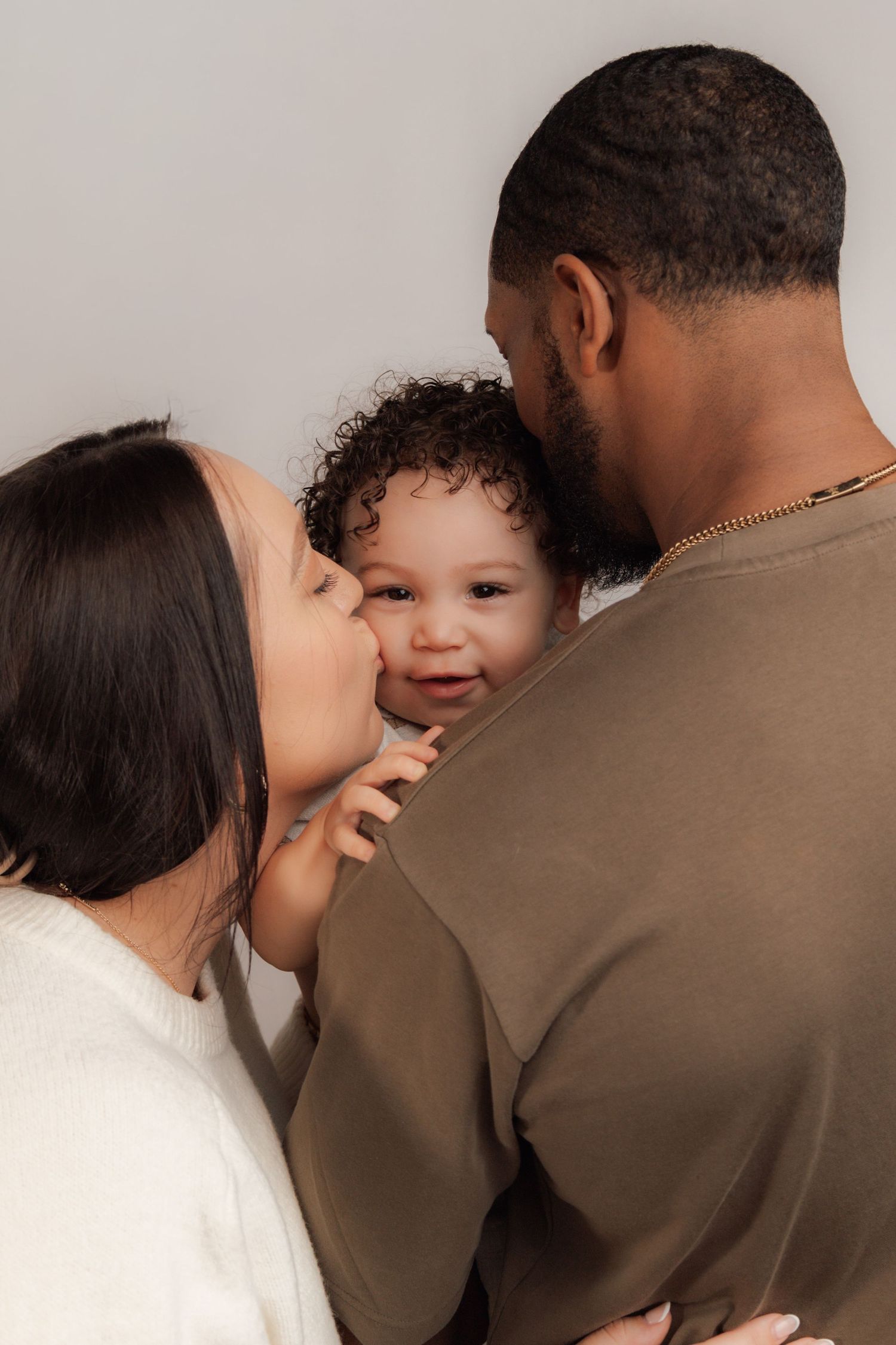 A loving family portrait with parents kissing their smiling curly-haired baby against a light background.