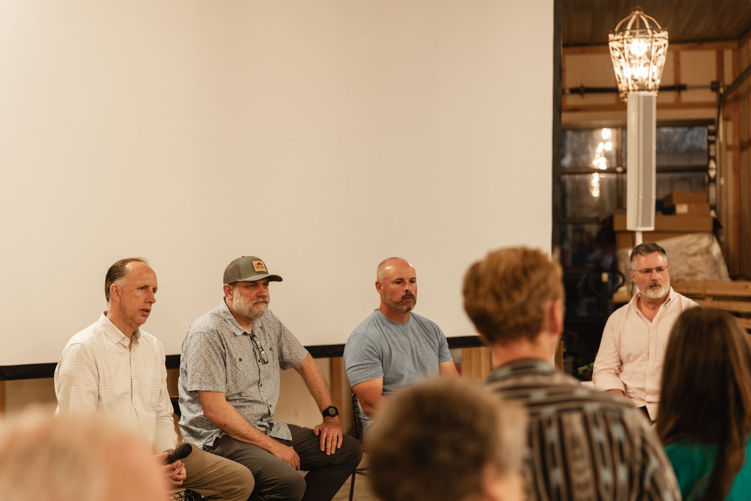 Four men sit in chairs during an indoor panel discussion in front of a white screen in a warmly lit venue.