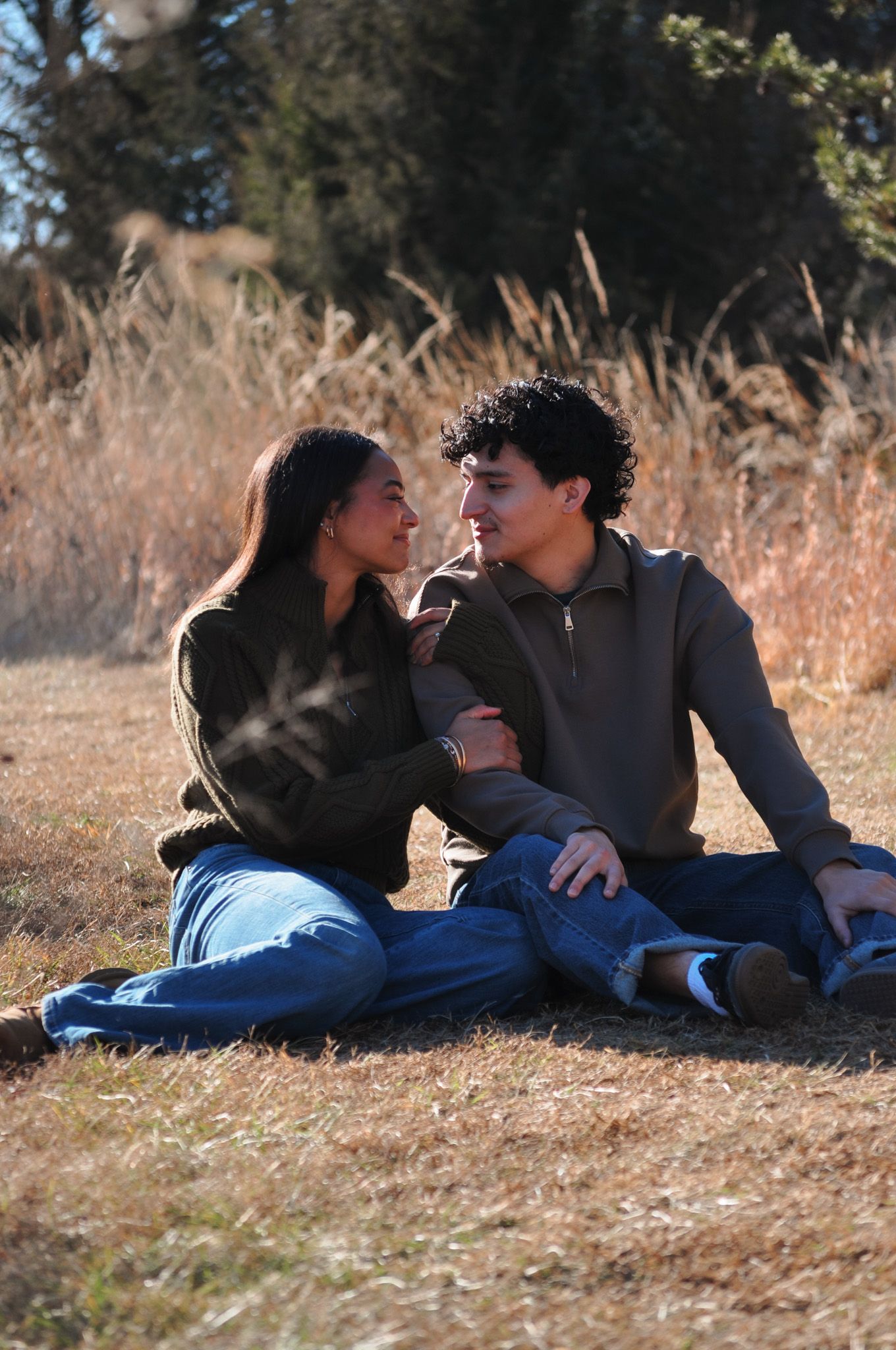 A young couple sits together on dry grass in a field, gazing lovingly at each other on a sunny day.