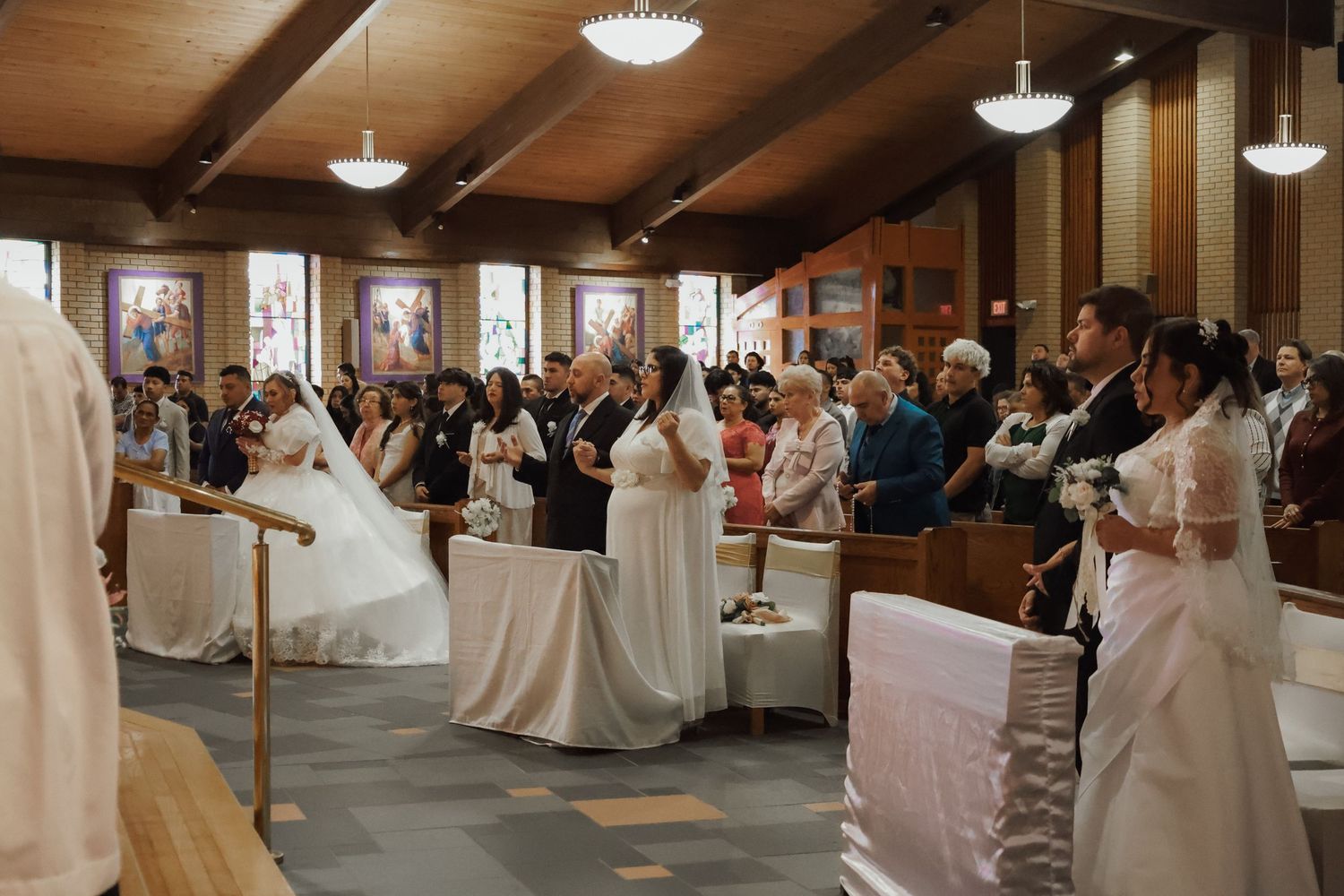 Multiple couples in white wedding attire stand at the altar during a group wedding ceremony inside a church.