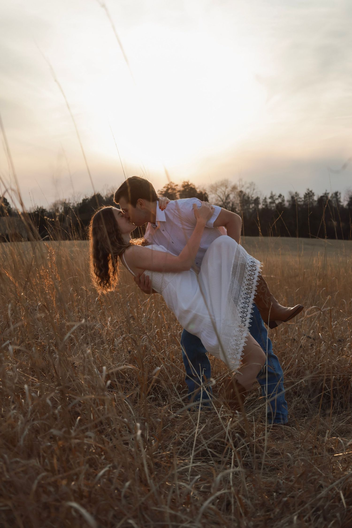 Couple sharing a romantic kiss in a golden field at sunset, man lifting woman in white dress and cowboy boots.