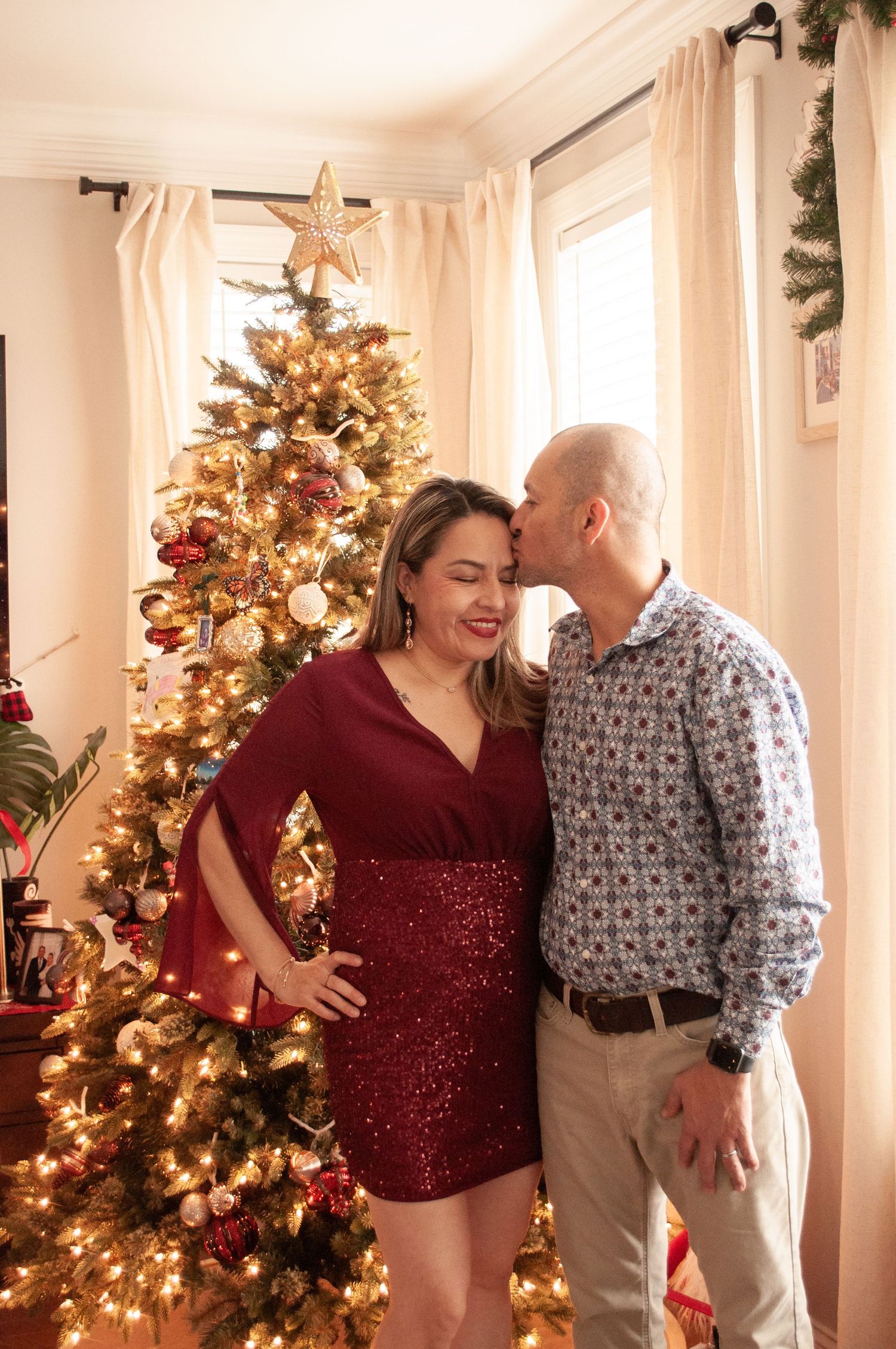 A couple poses by a decorated Christmas tree, the man kissing the woman's cheek as she smiles in a red sequin dress.